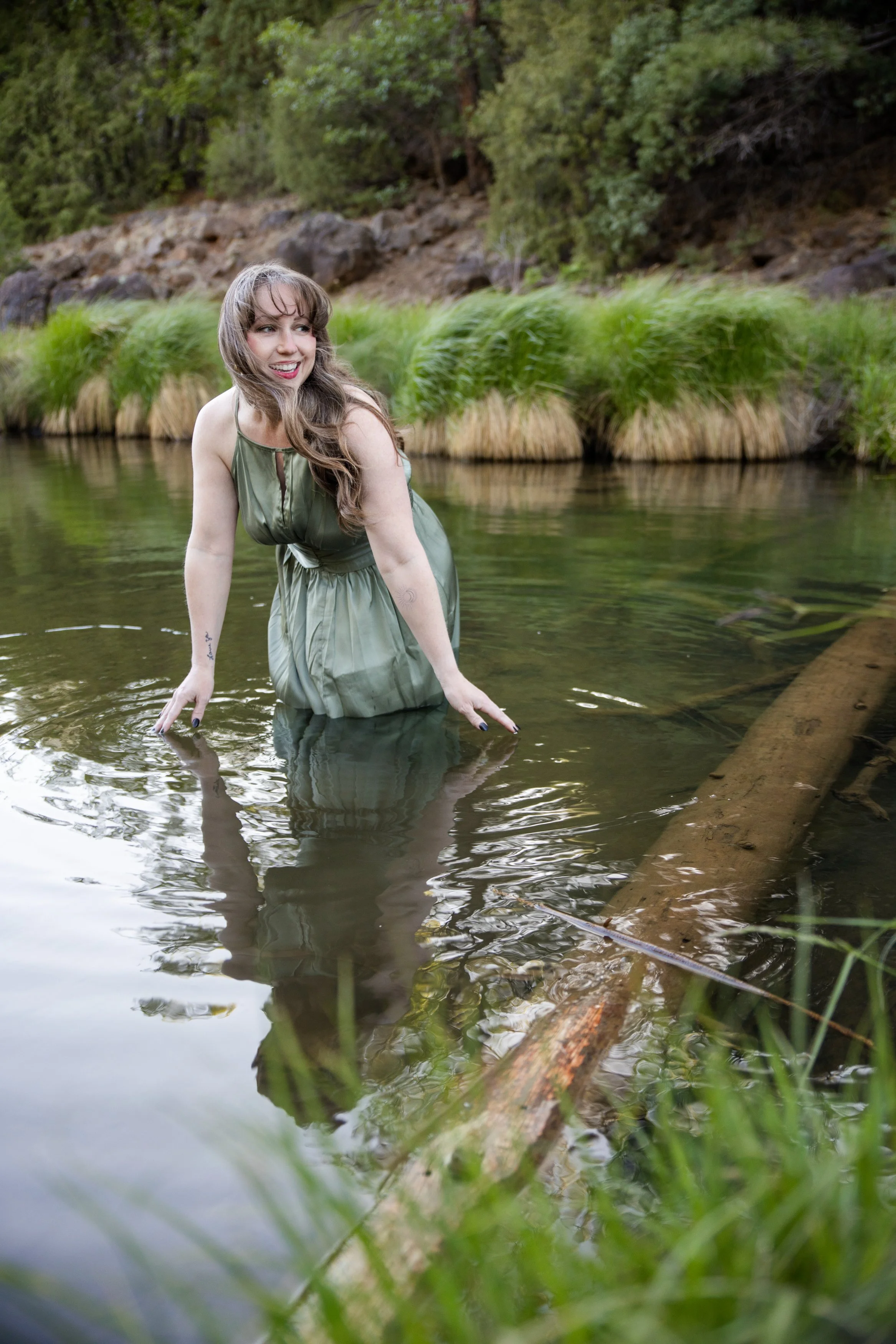 Woman in a green dress standing in a river surrounded by greenery and trees.