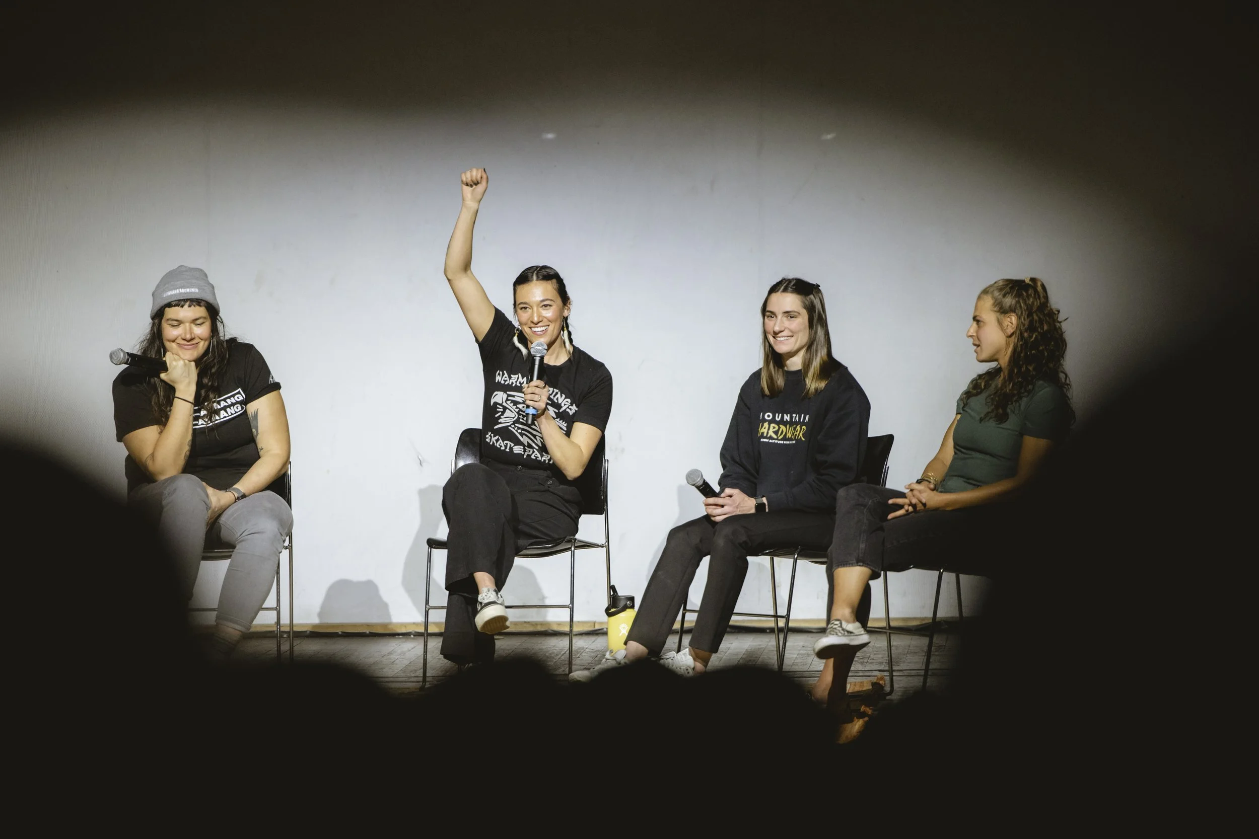 Panel of four women seated on stage, two holding microphones, one raising her hand, in a well-lit room with a plain white wall background.