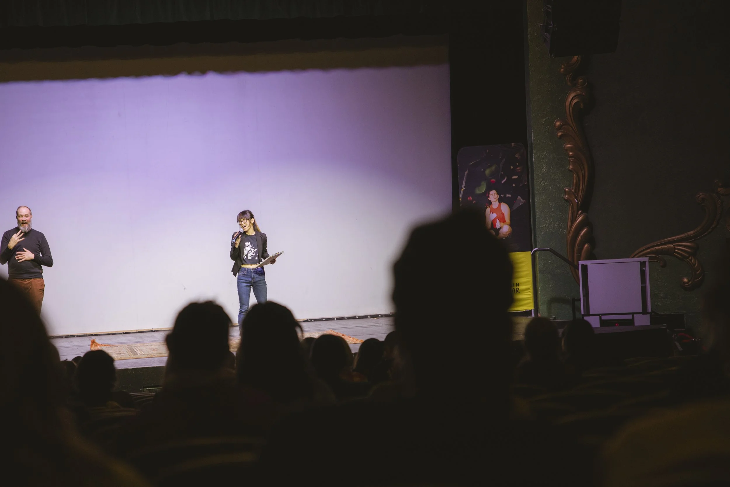 People on stage during a presentation or performance, with audience in foreground, large plain backdrop, and decorative wall details to the right.