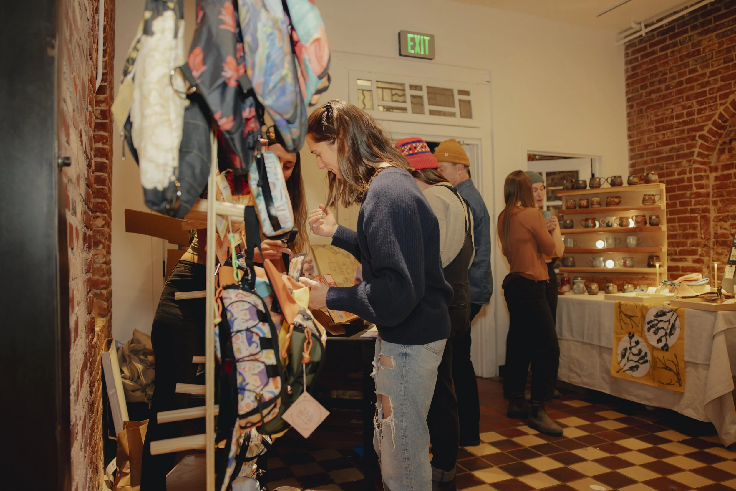 People browsing handcrafted items at an indoor craft market or boutique shop, with shelves of pottery and decorative items in the background.