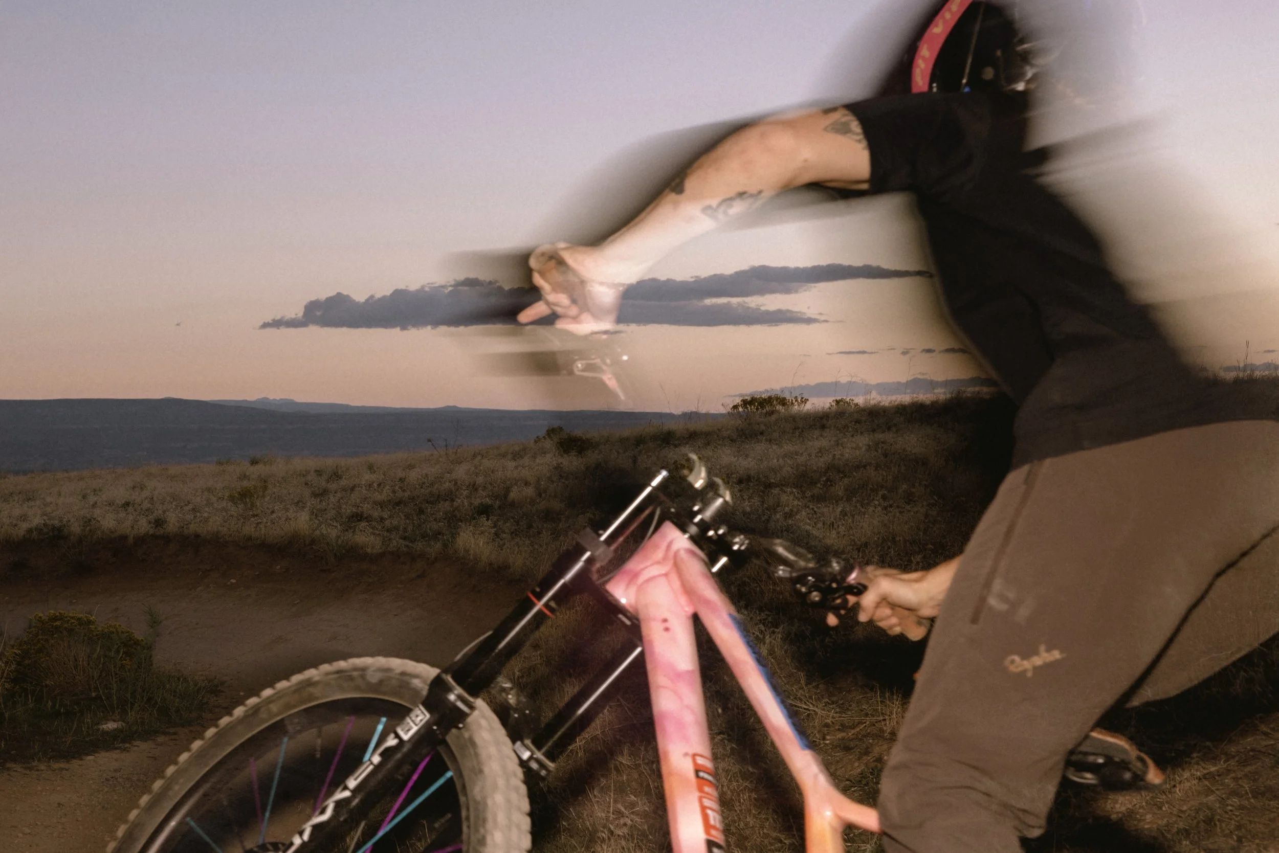 A mountain biker is riding on a trail during sunset with a mountainous landscape in the background. The rider is wearing dark clothing and is holding the bike's handlebars.