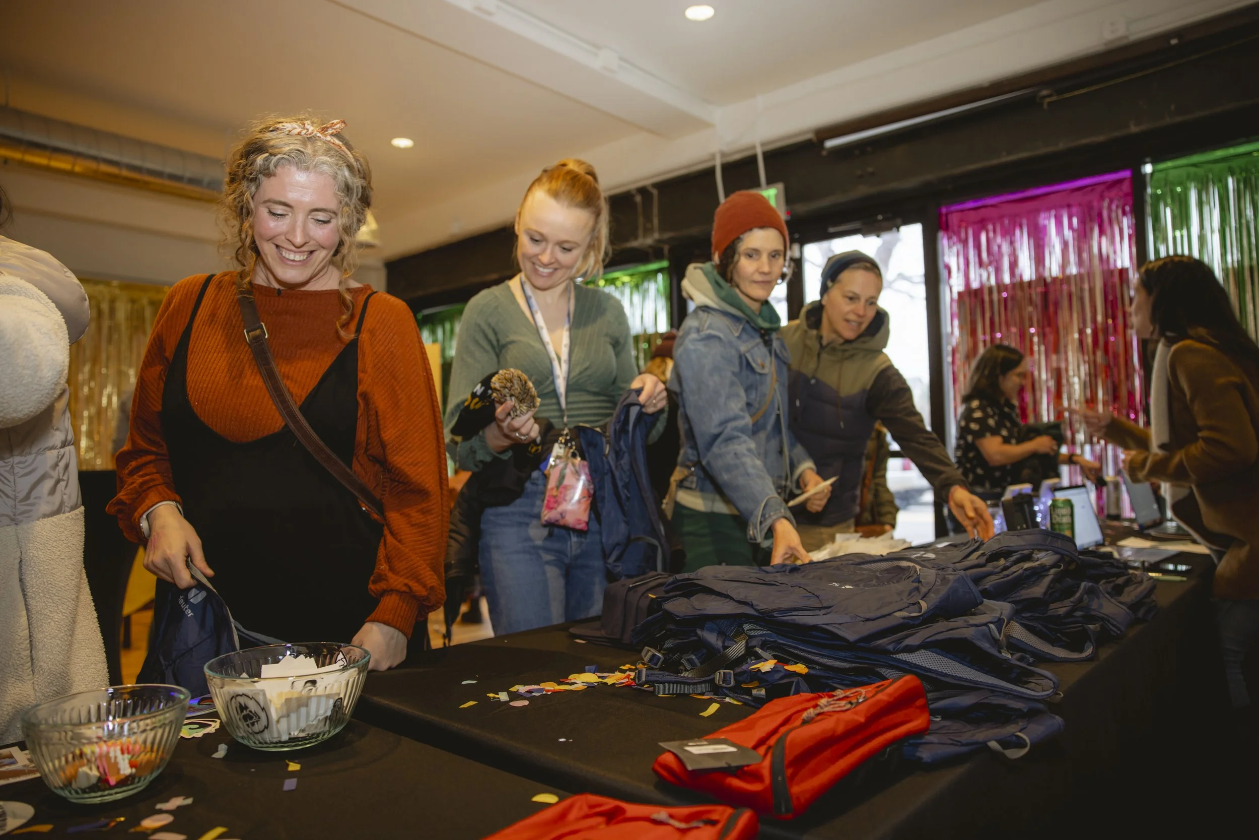 A group of women shopping at a registration or check-in table at an indoor event, with backpacks, hats, and other items on the table, and colorful curtains in the background.