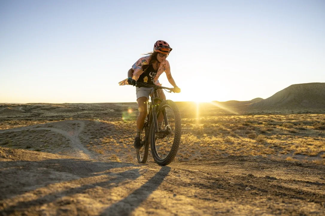 A woman riding a mountain bike on a dirt trail in a desert landscape during sunset, wearing a helmet and sunglasses.