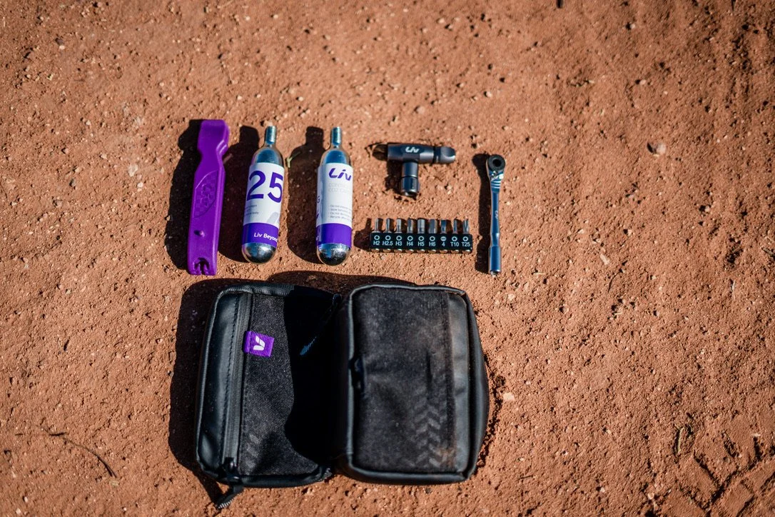 A black pouch and tools and supplies for tire maintenance on a dirt surface, including two CO2 cartridges, a purple tire lever, a T-shaped valve tool, a hydraulic caliper piston tool, a small set of screwdriver bits, and a socket wrench adapter.