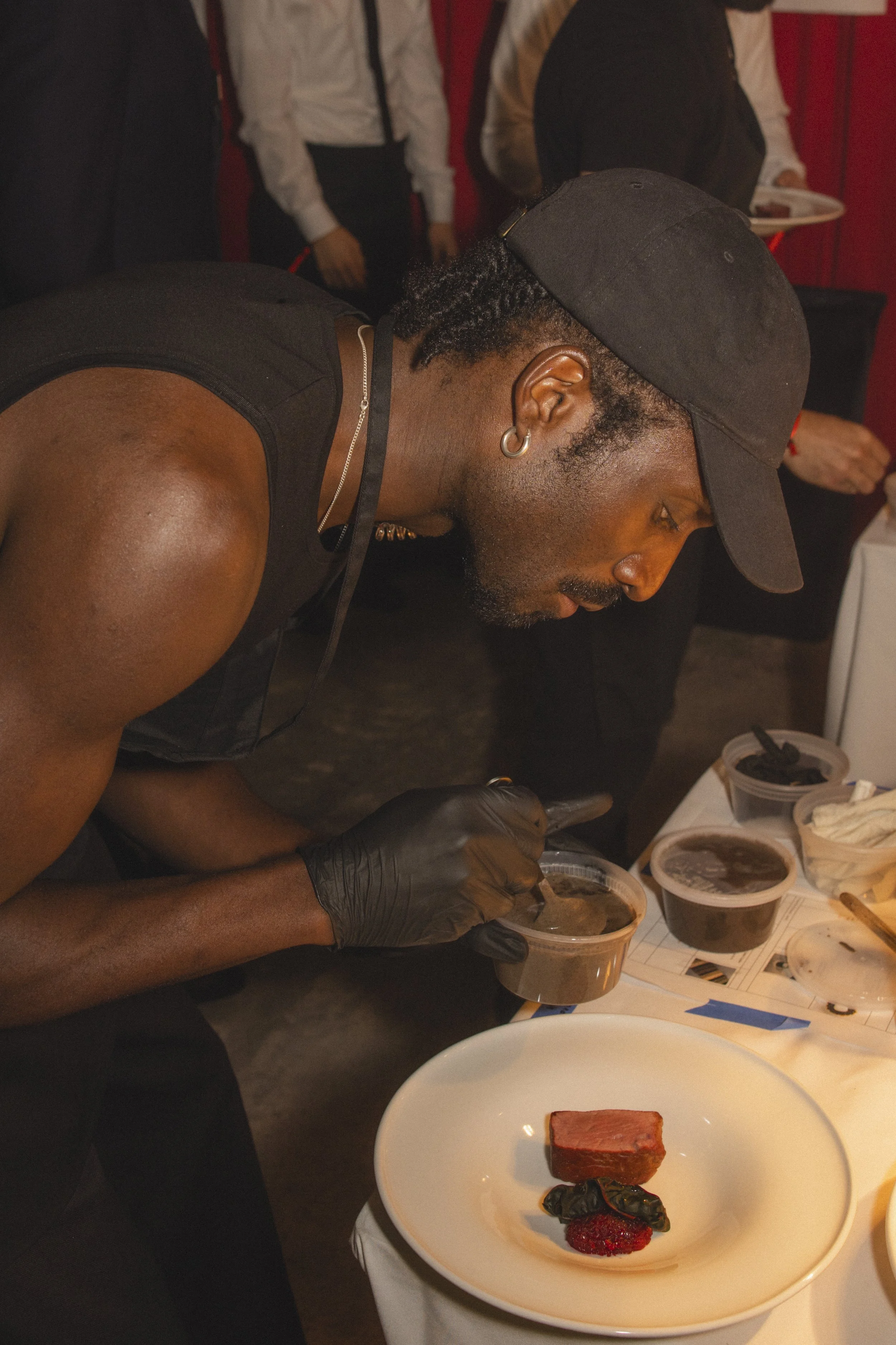 A man wearing a black cap, black gloves, and a sleeveless shirt is intensely examining or tasting a dish in a professional setting, with various dishes and utensils on the table.