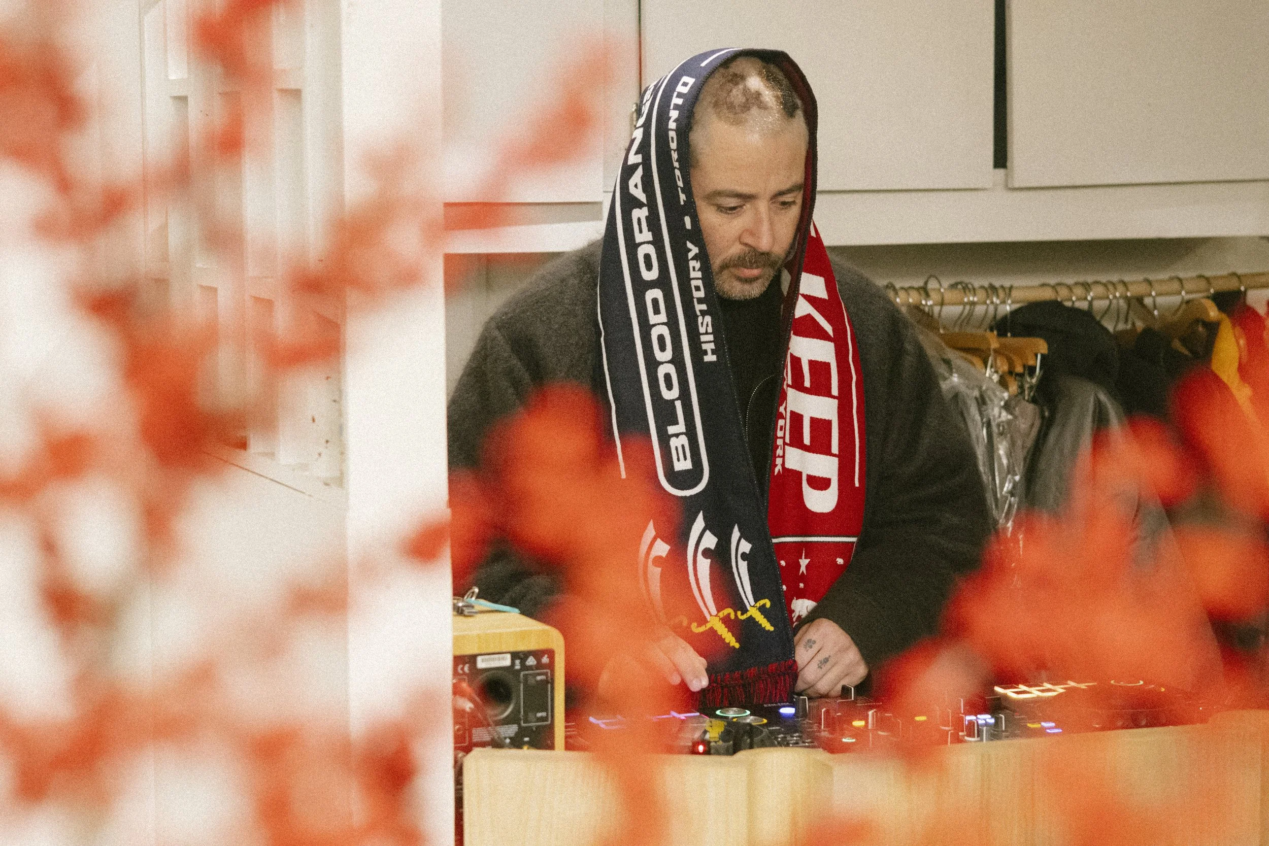 A man with a scarf on his head, wearing a dark sweater, is DJing at a table with DJ equipment in a room with clothing racks. The photo is framed with orange flowers in the foreground.