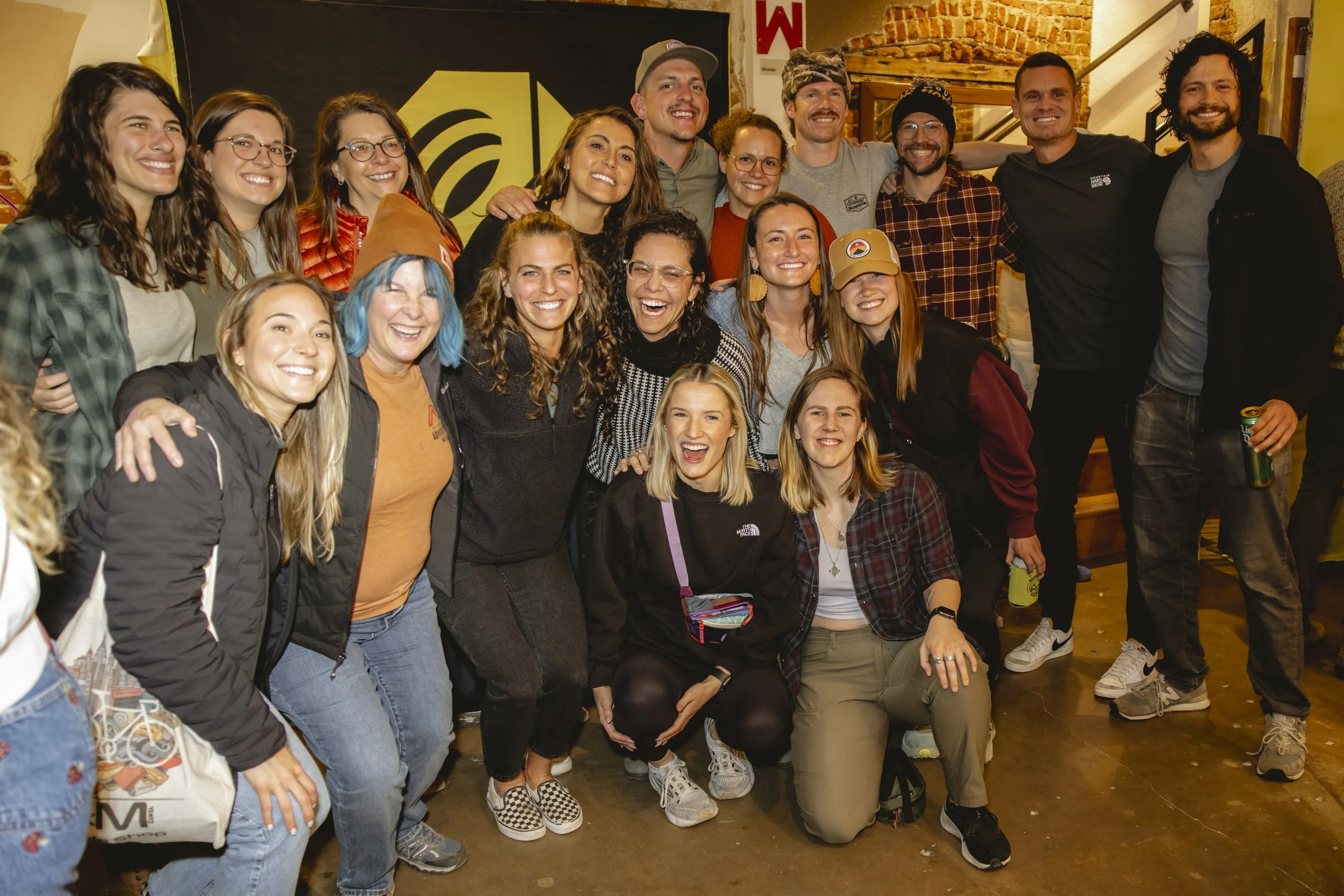 Group of smiling people, mostly women, indoors at a social gathering, standing close together, some with arms around each other, in front of a black and yellow background.