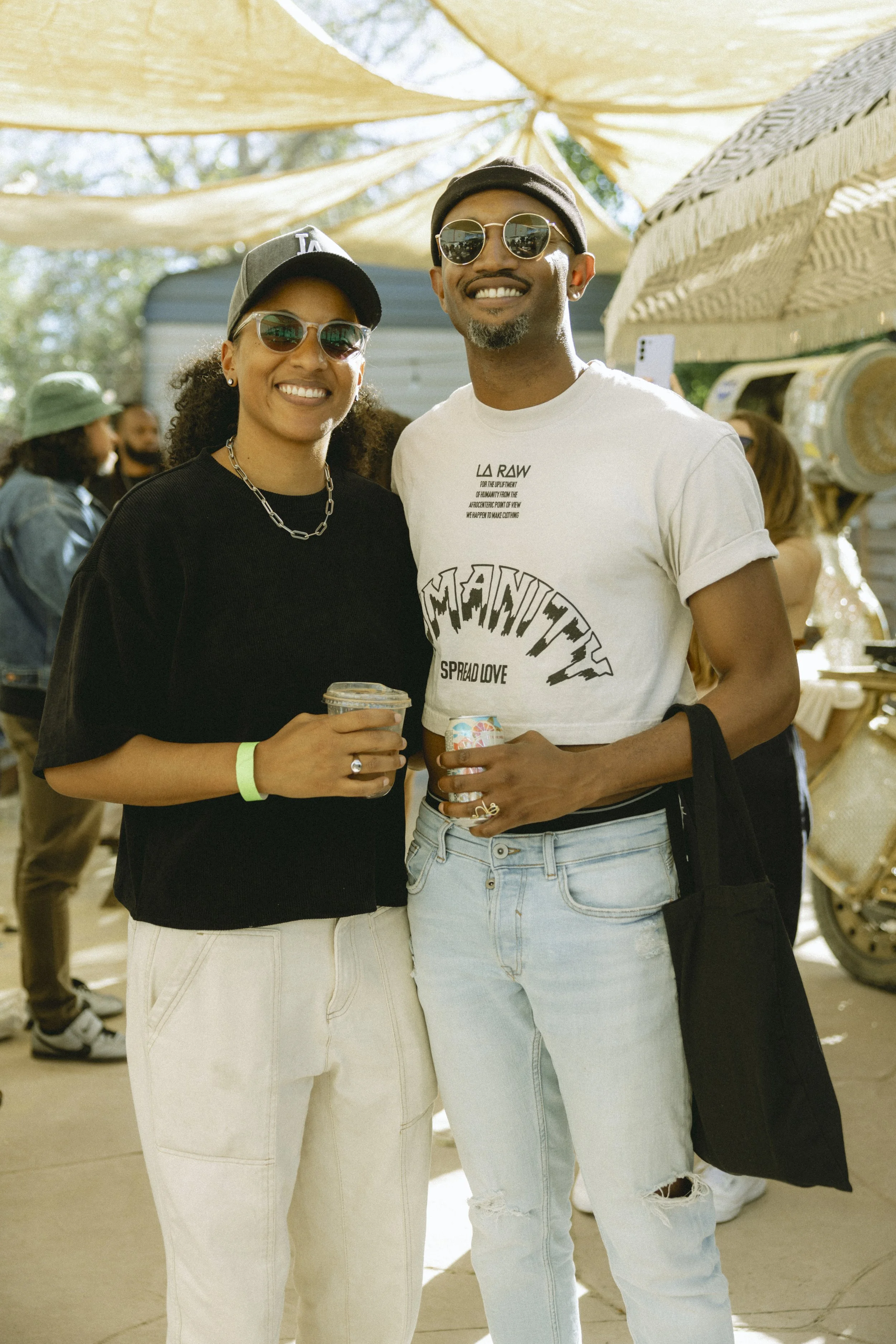 A smiling couple at an outdoor event, both wearing sunglasses and casual clothing, holding beverages, with other people and decorated tents in the background.