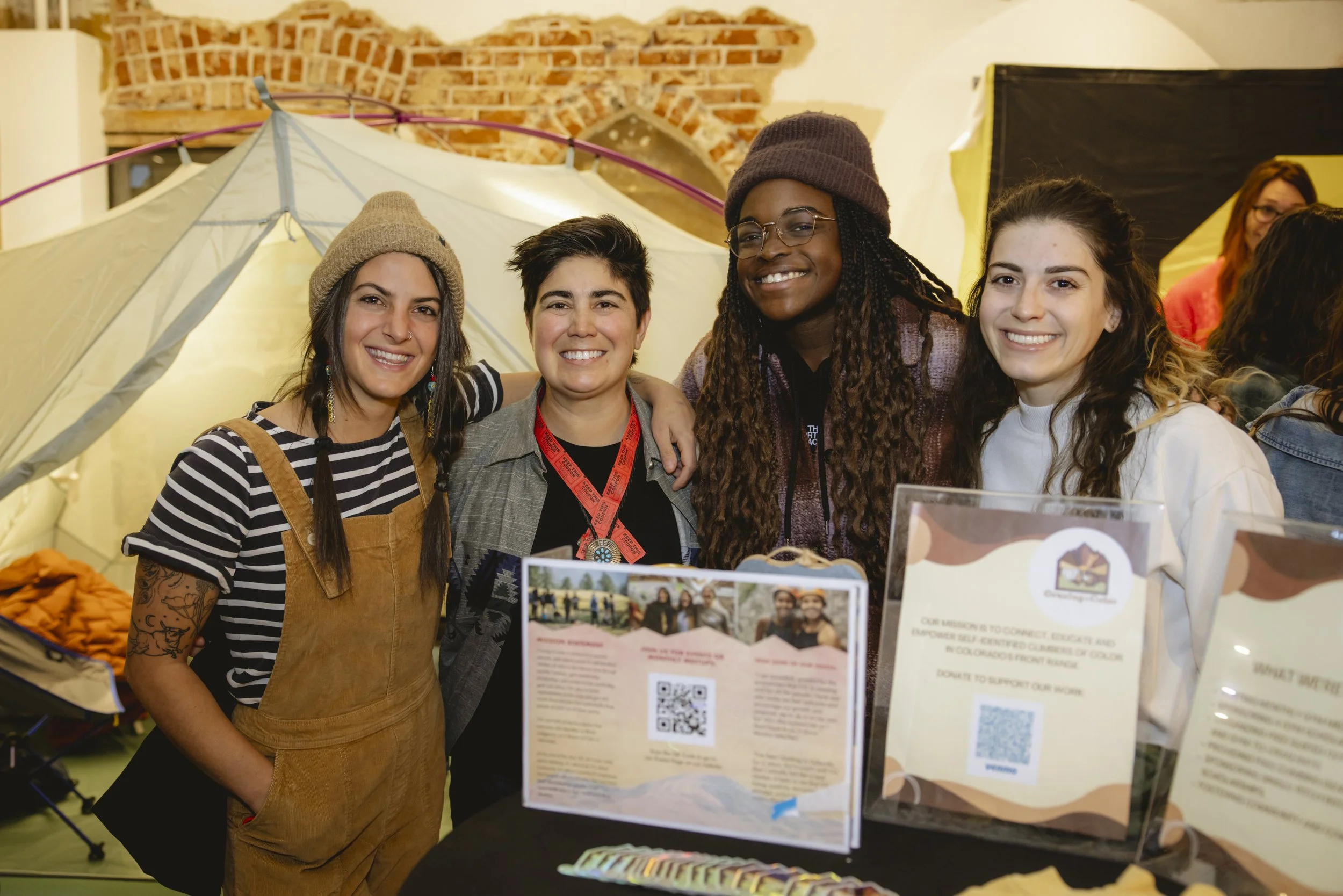 Four women smiling and standing close together at an indoor event, with informational display boards and a tent in the background.