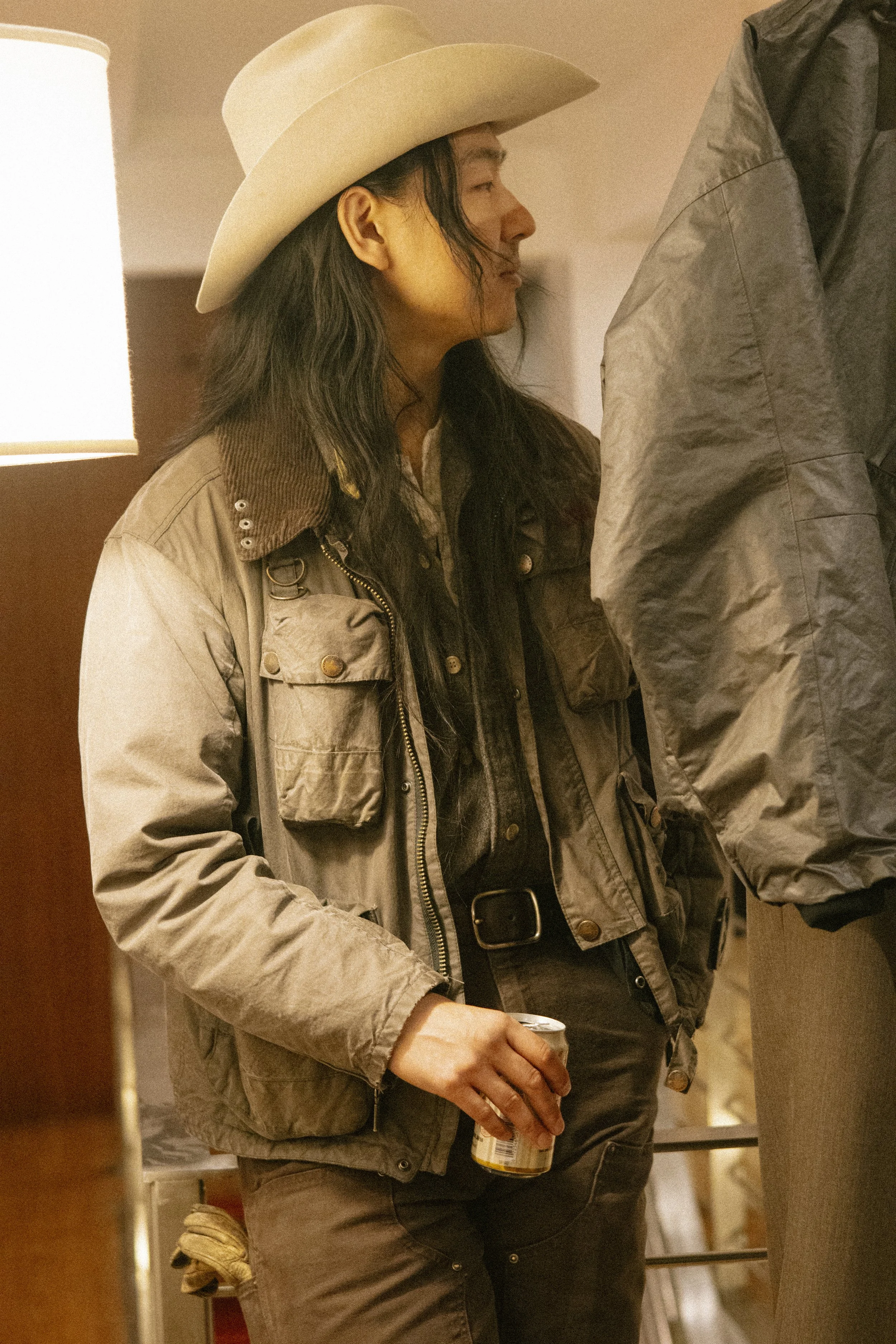 A woman with long hair wearing a beige cowboy hat, a brown jacket, and holding a can in her hand, standing inside a room with beige walls and a lamp in the background.