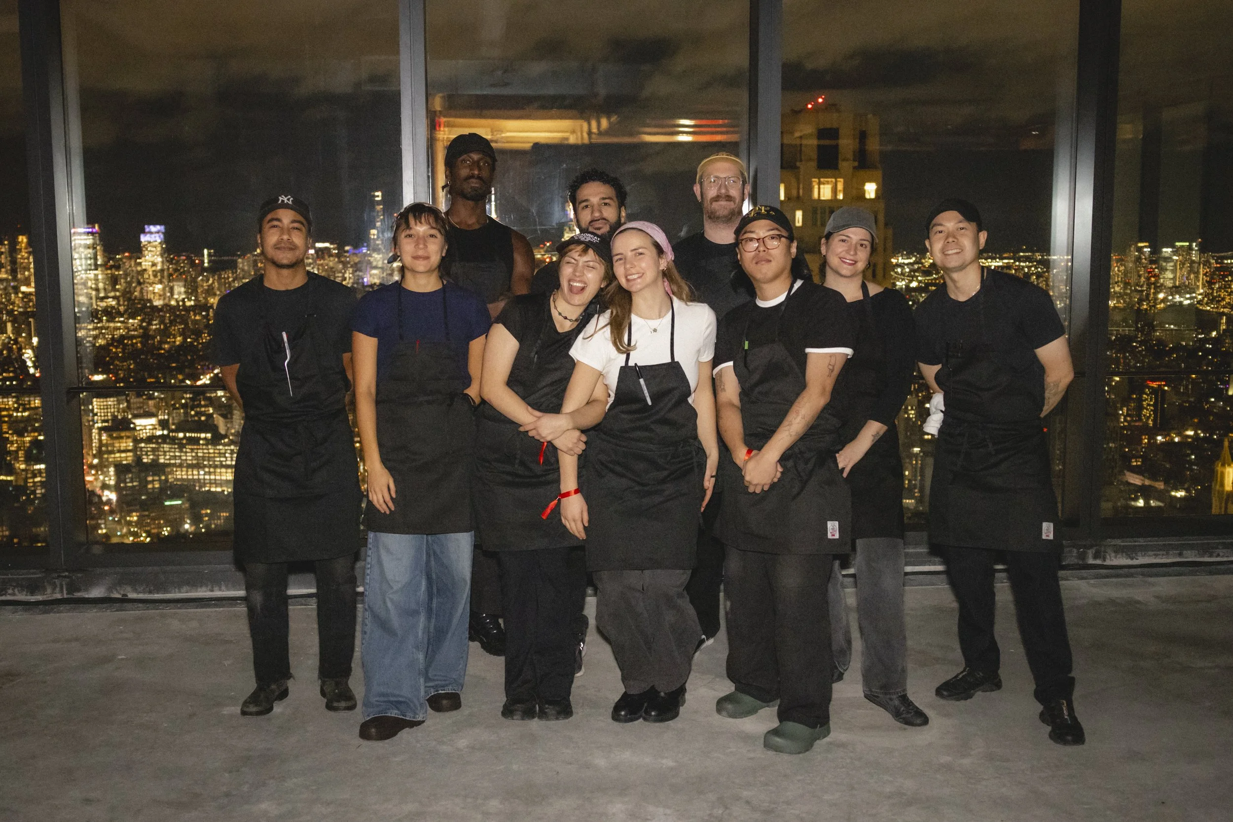A group of ten people, including men and women, standing on a rooftop at night with a city skyline illuminated in the background. They are dressed in casual black outfits, some wearing aprons and hats, likely indicating they are restaurant staff or c