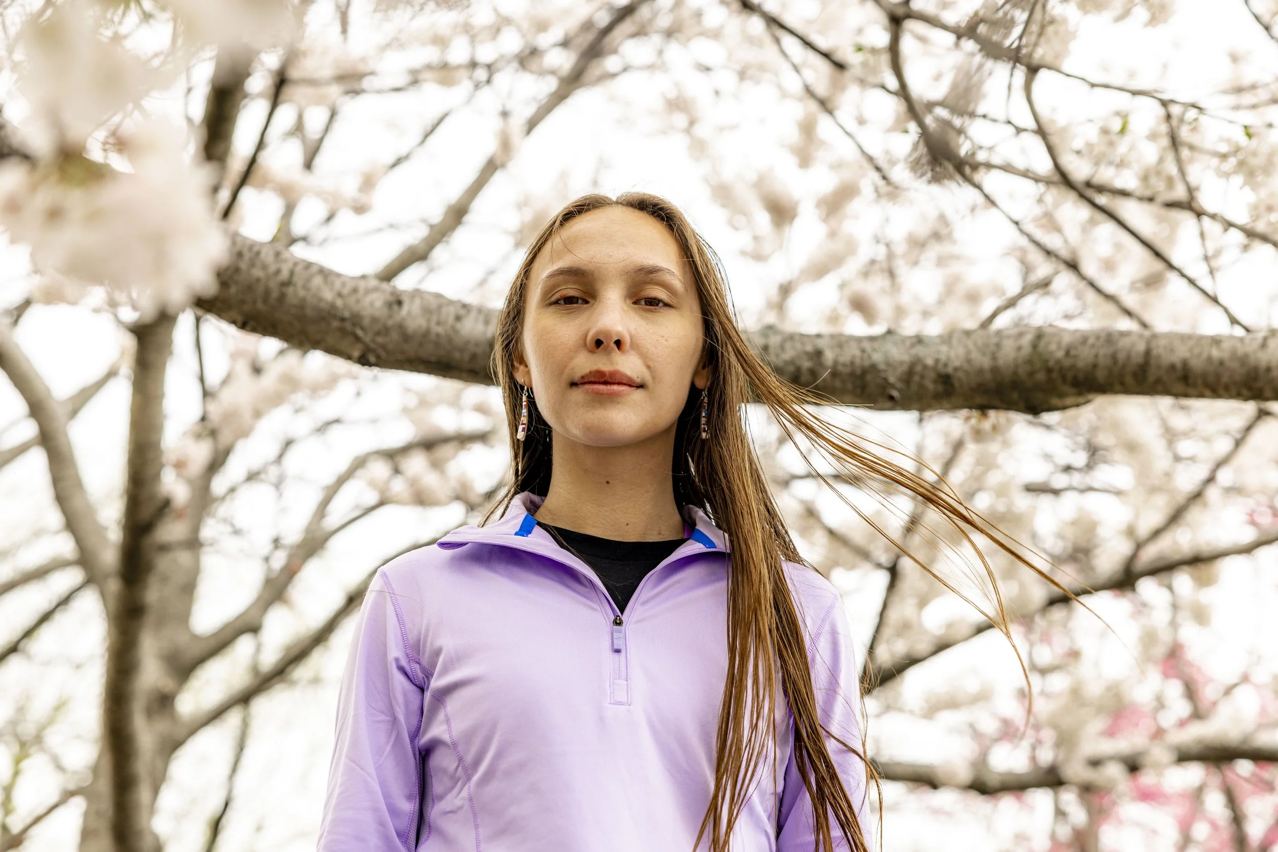 A woman with long brown hair and earrings standing under cherry blossom trees, wearing a purple athletic jacket.