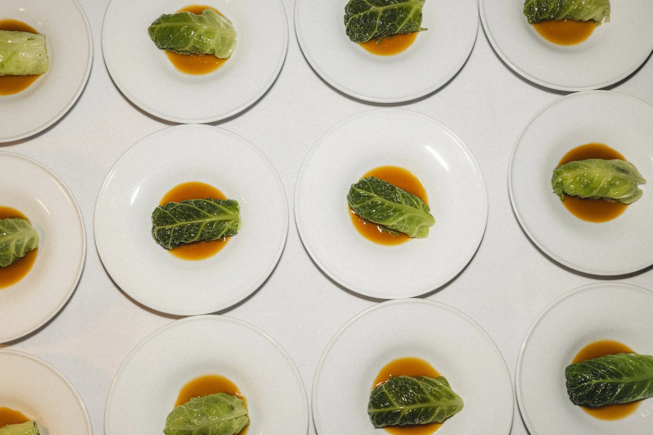 Multiple white plates, each with a stuffed green cabbage leaf on a bed of orange sauce, arranged in a grid pattern.
