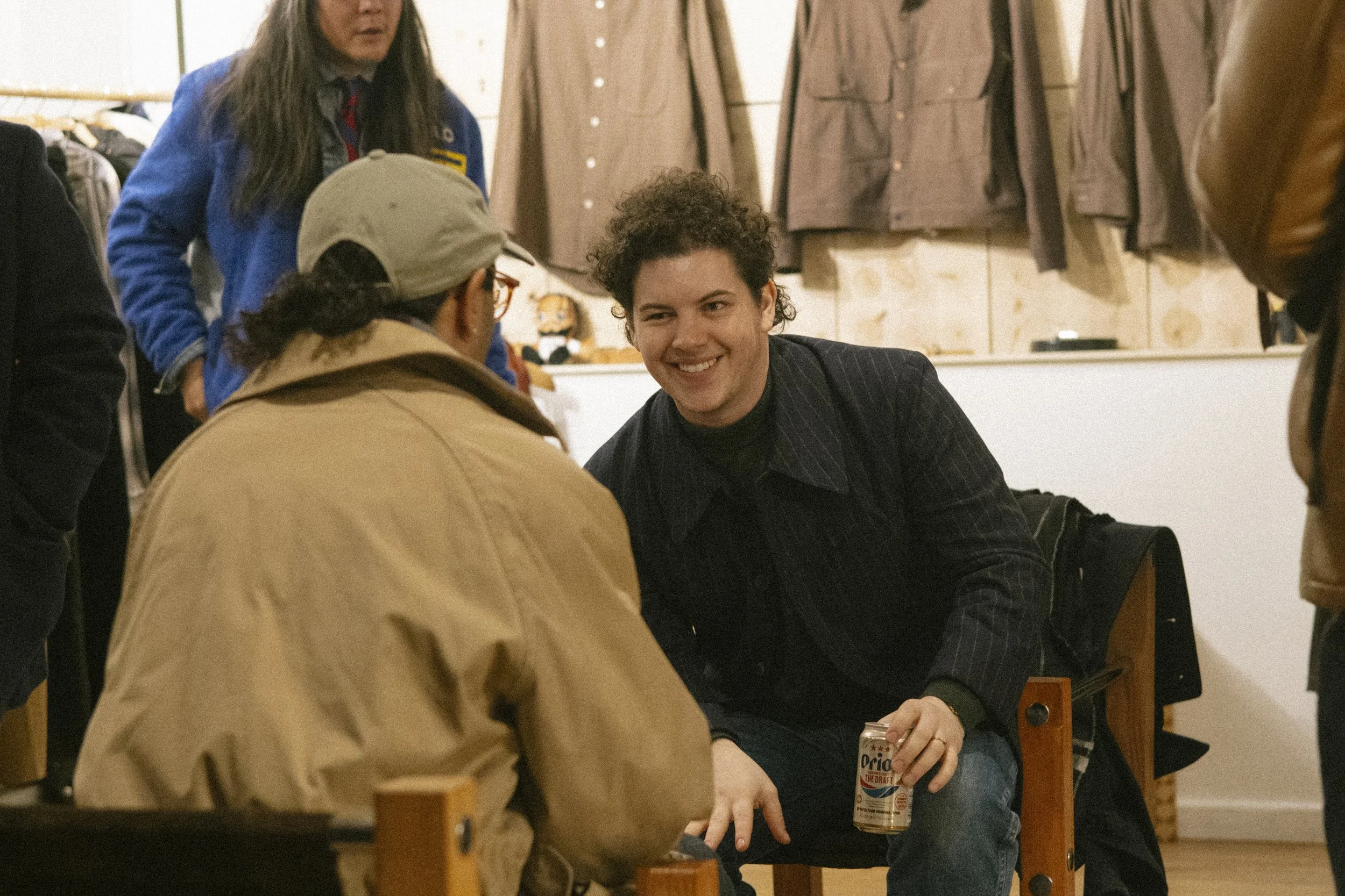 A man with curly hair smiling and holding a beer can, sitting in a store or cafe, talking to a person with a baseball cap. Others are standing nearby, and jackets are hanging on the wall in the background.