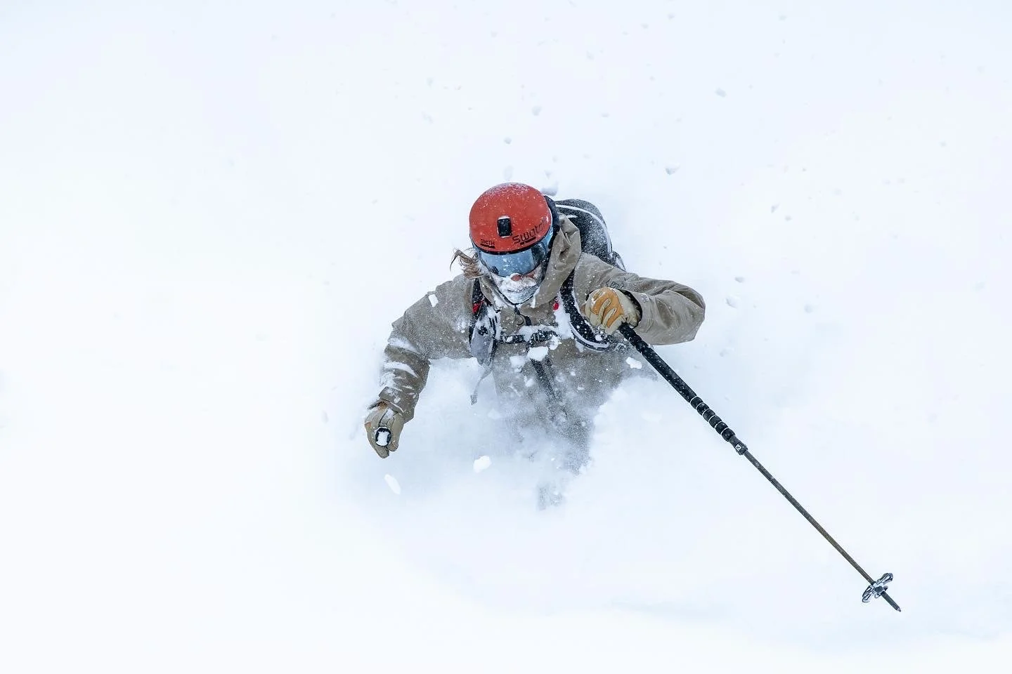 Person in beige jacket, red helmet, and goggles climbing snowy mountain with trekking pole