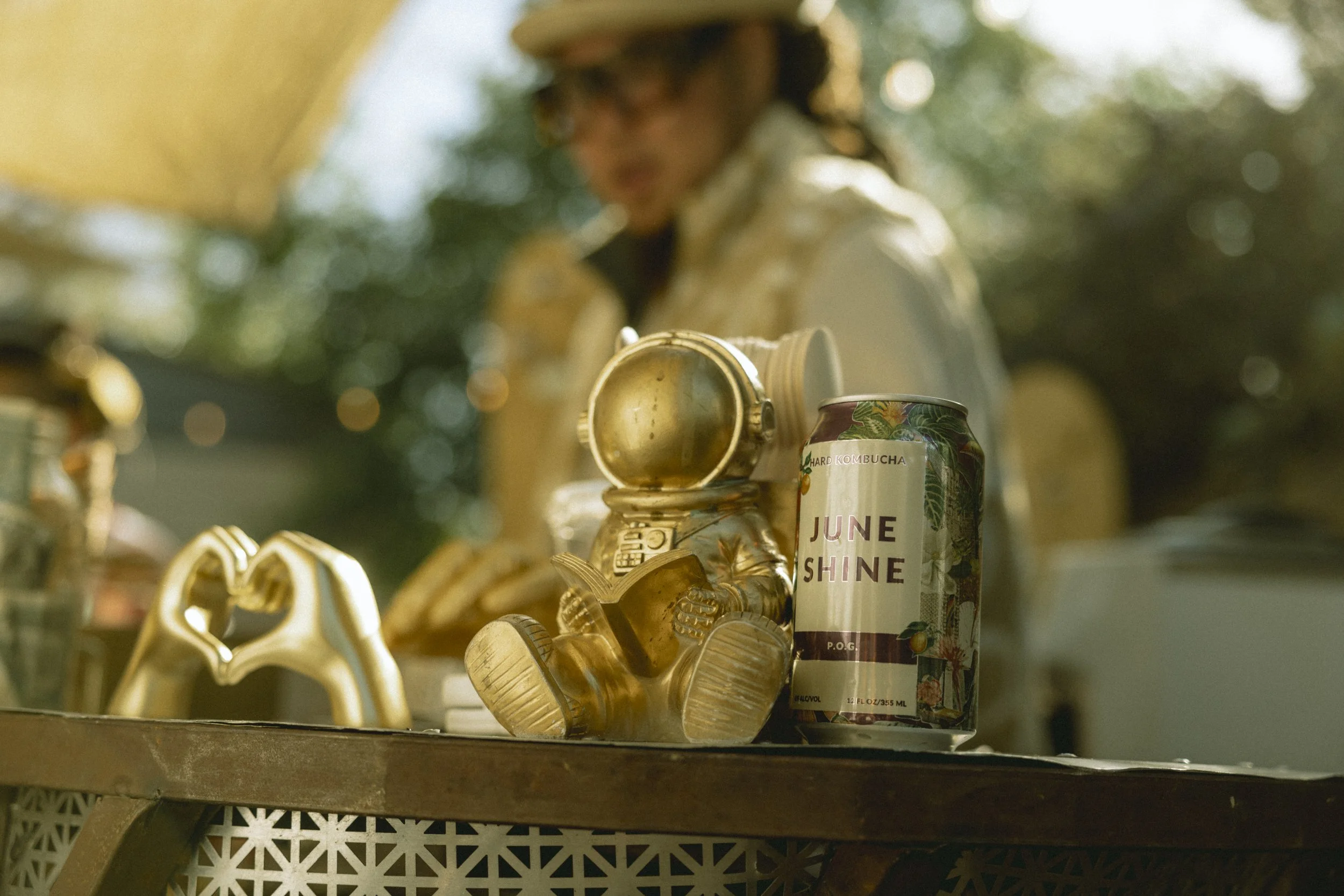 A gold-colored astronaut figurine sitting on a wooden surface next to a can labeled 'June Shine' and a person's blurred face in the background.