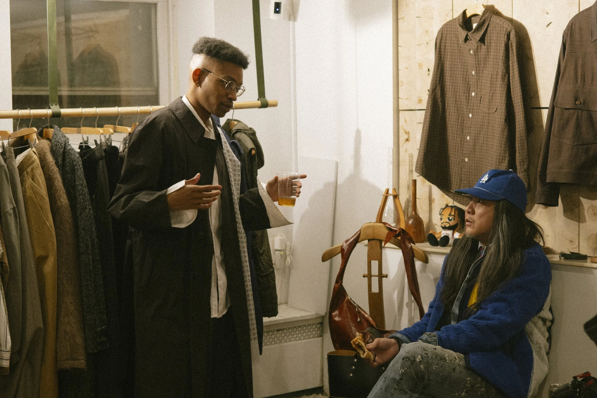 Two people in a vintage clothing store having a conversation. One person is standing, holding a drink, and the other person is sitting, wearing a blue baseball cap and holding a sandwich.
