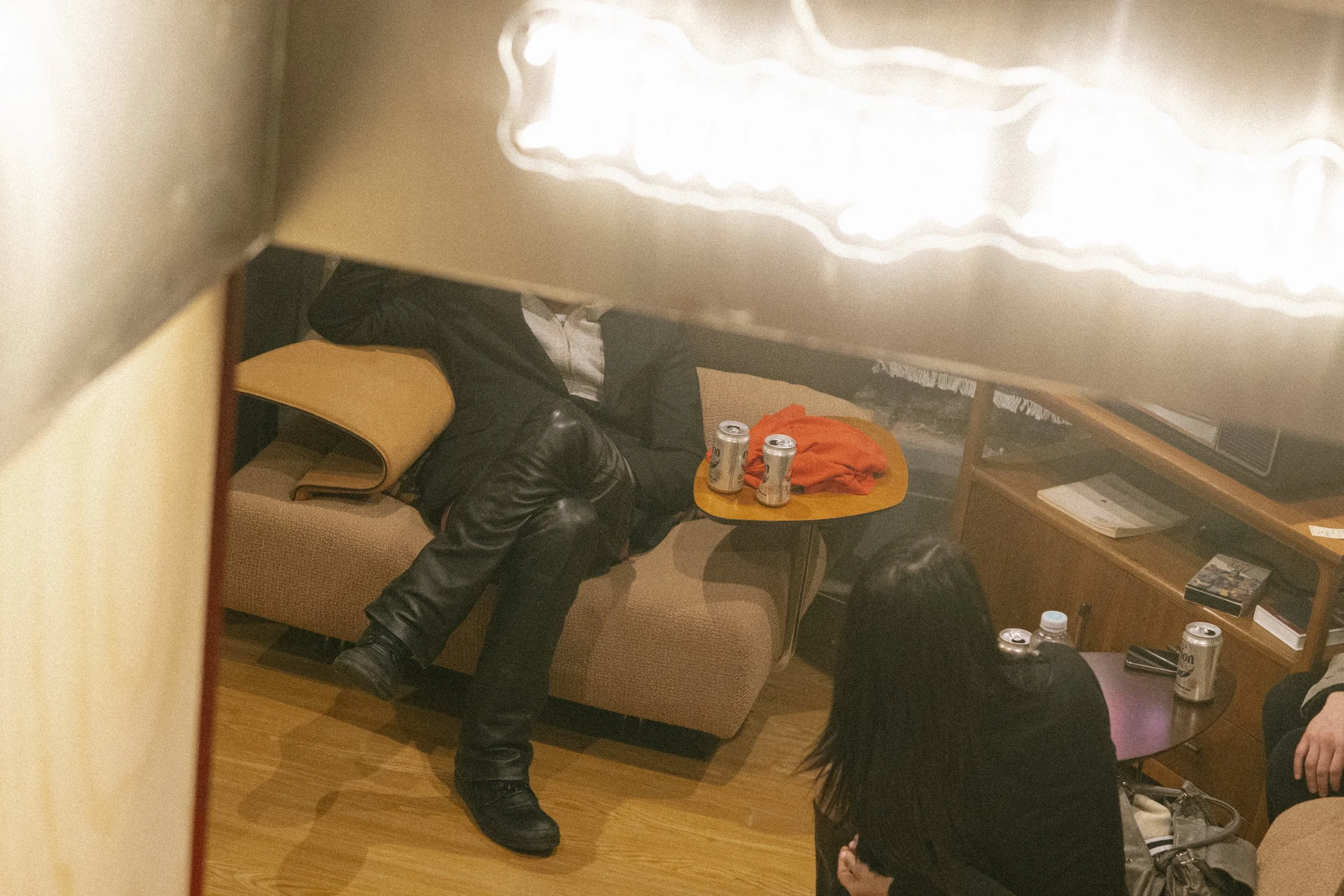 A reflection in a mirror showing a man sitting on a couch with a woman sitting nearby, and a small table with two cans of drink and a bag, in a room with wood furnishings.