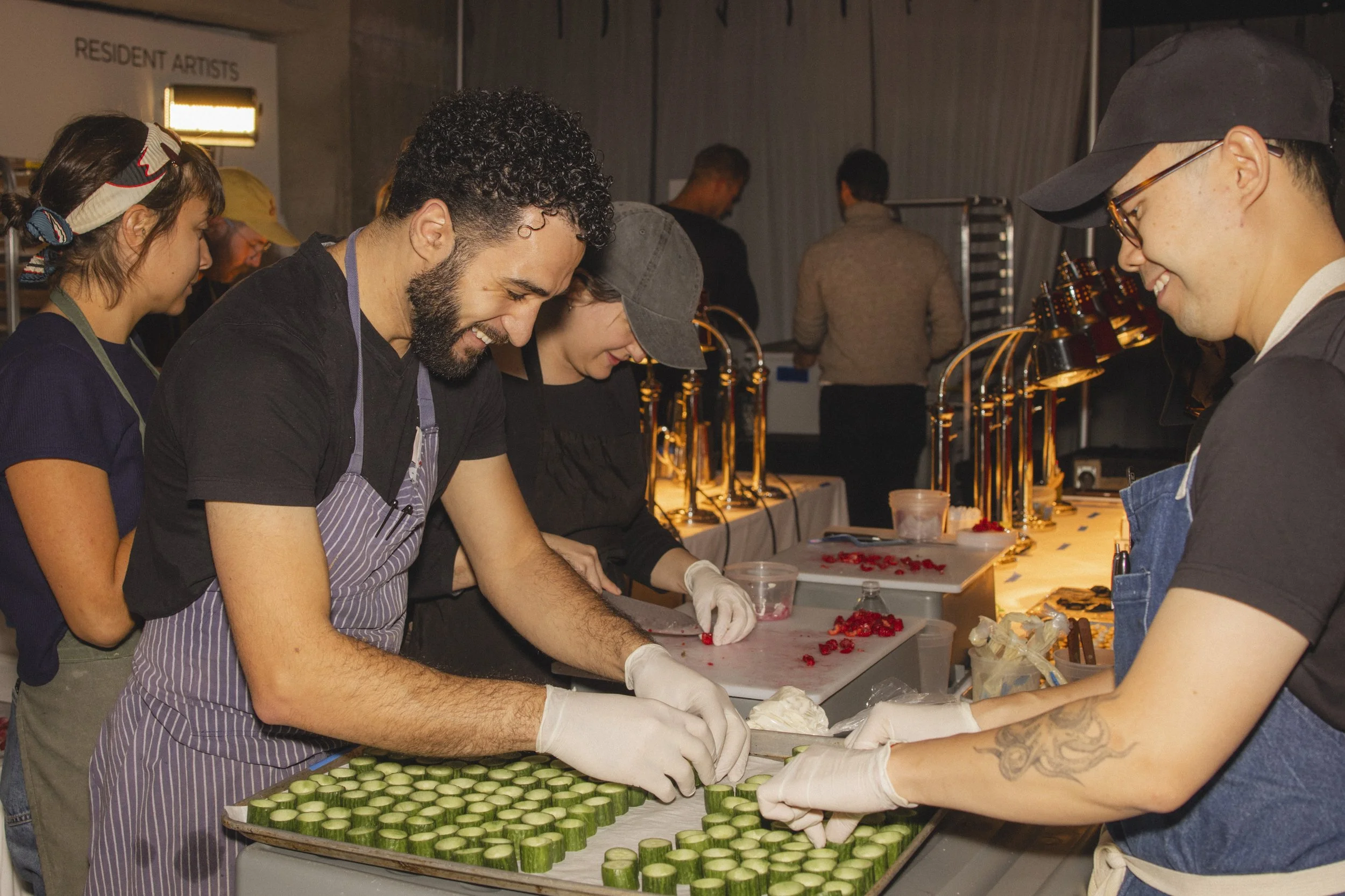 People at a food station preparing sliced cucumbers and strawberries, wearing gloves and aprons, smiling and enjoying their work.