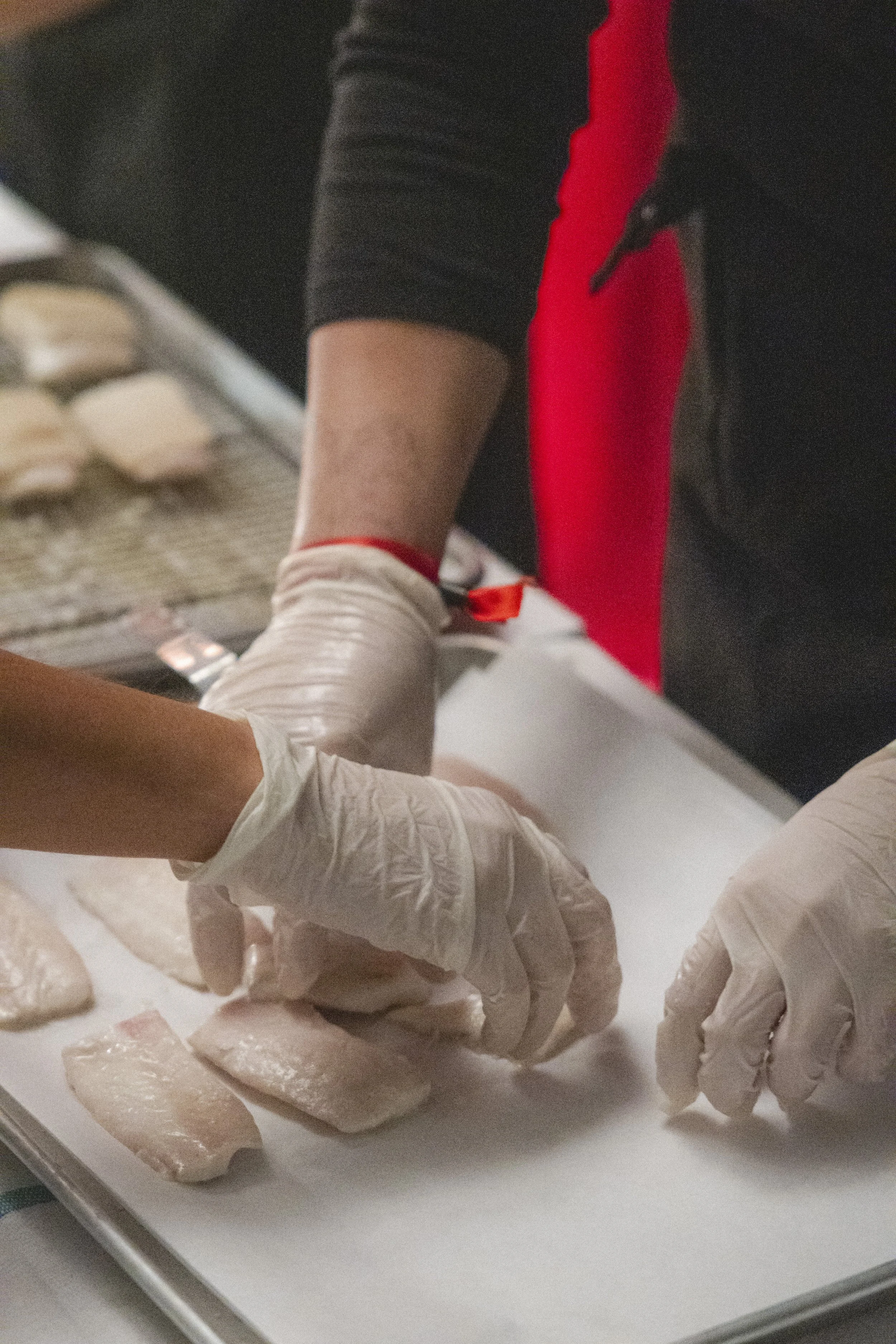 People wearing gloves preparing chicken fillets on a white cutting board in a kitchen.