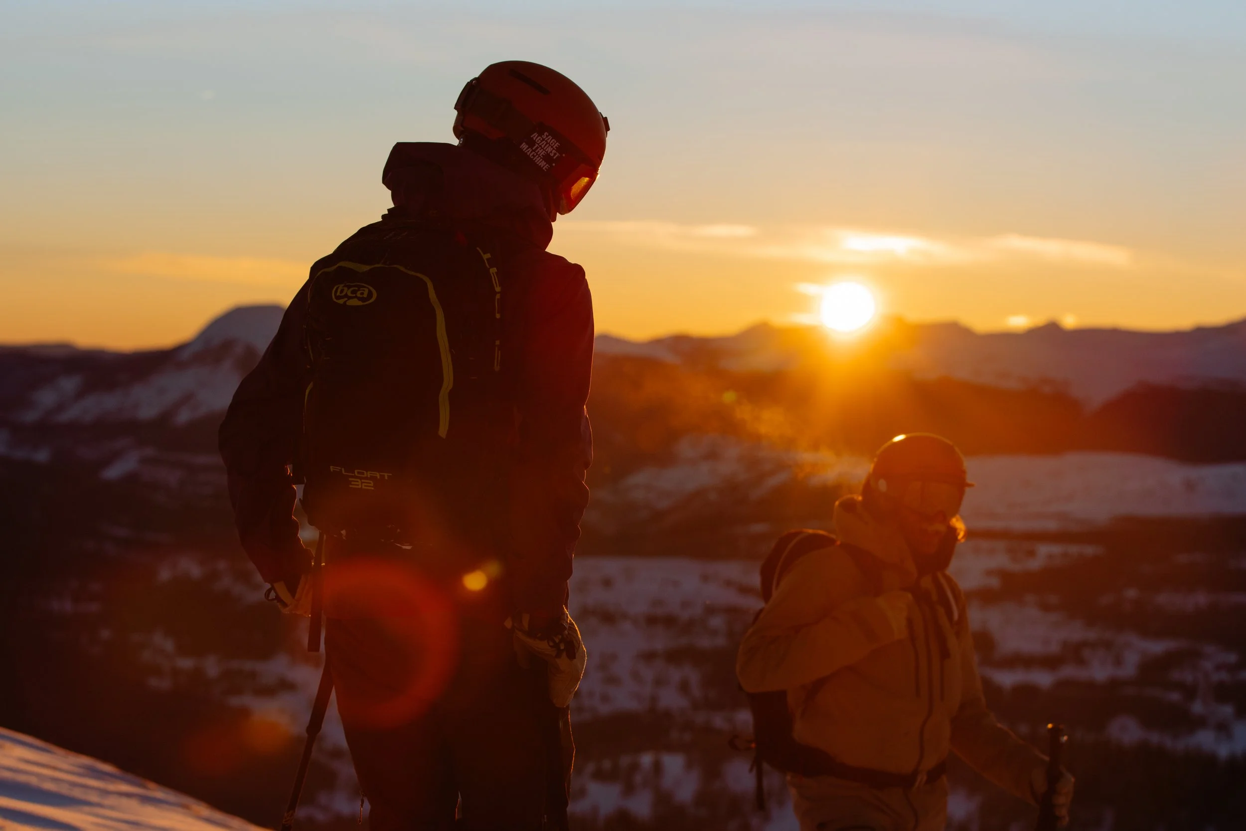 Two skiers in winter gear and helmets standing on snowy mountain during sunset with snow-covered mountains and a colorful sky in the background.