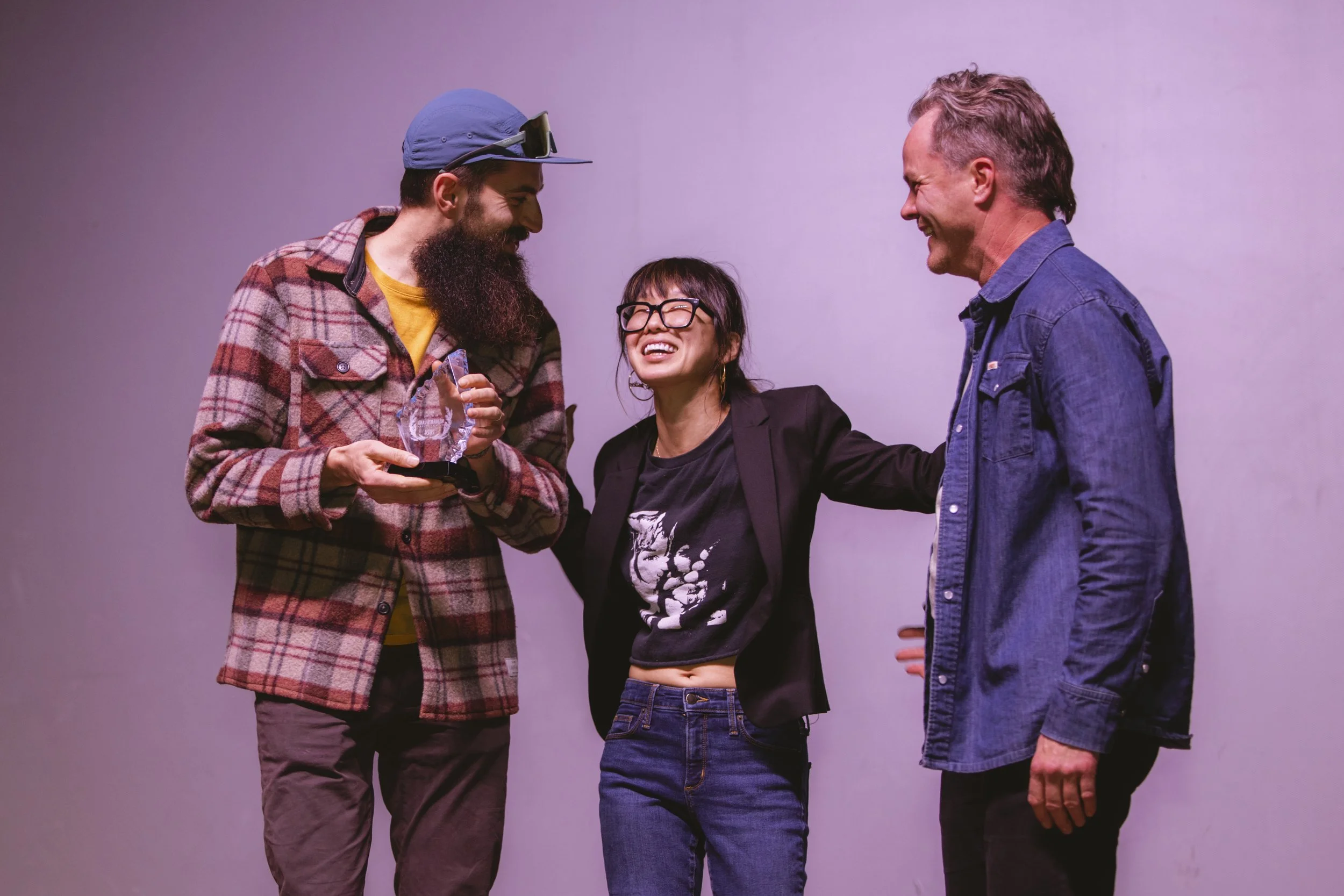 Three people laughing and celebrating. One person is holding an award, another has their arm around the first, and the third is smiling.