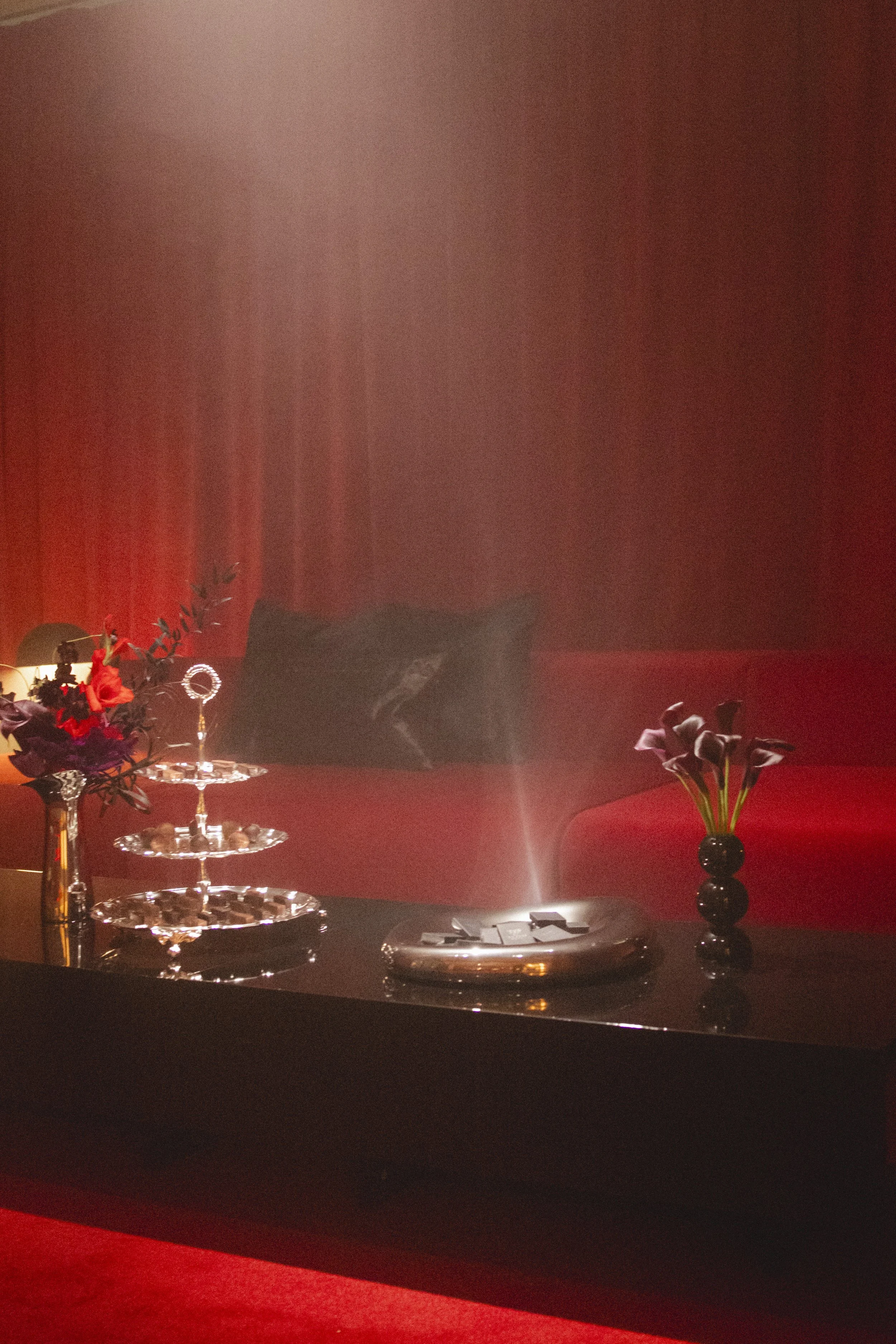 A living room with dark decor, featuring a red sofa, a black side table with purple calla lilies in a black vase, a bouquet with red and purple flowers in a glittering glass vase, a silver tray with small items, and a soft mist or smoke in the air.
