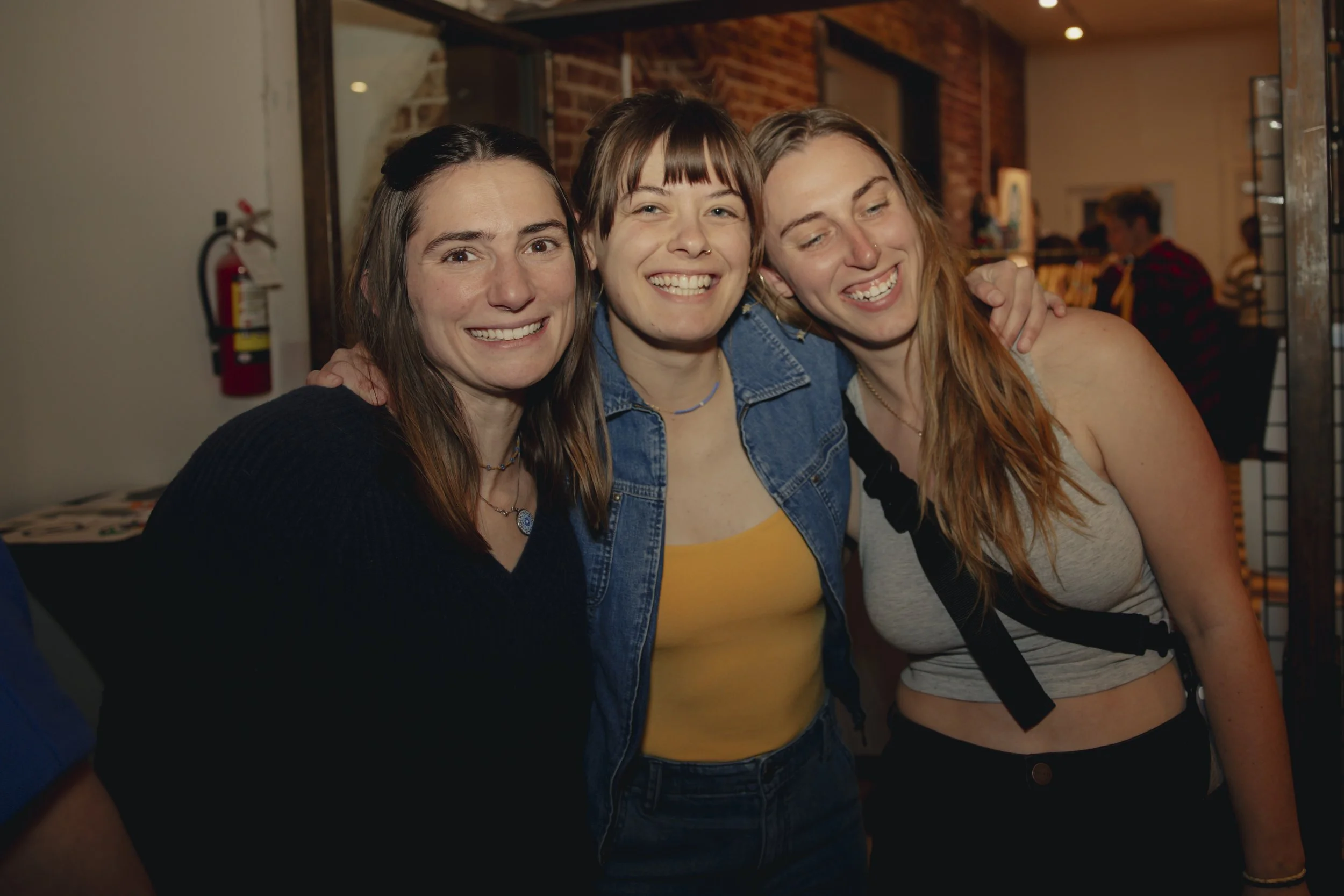 Three young women smiling and posing together at an indoor social gathering.