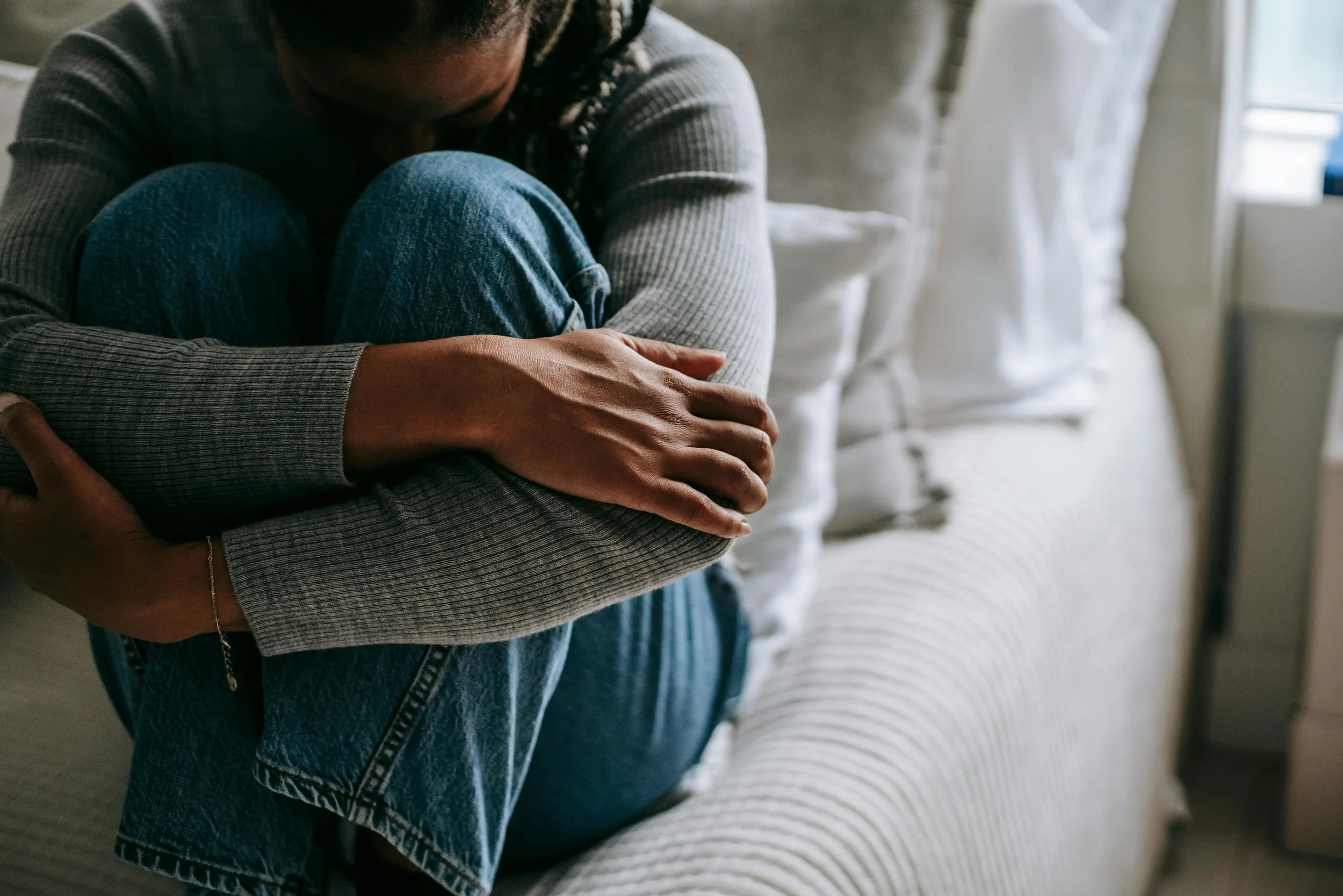 A person sitting on a bed with knees pulled to their chest, arms wrapped around themselves, conveying emotional distress or withdrawal in a quiet, intimate space.