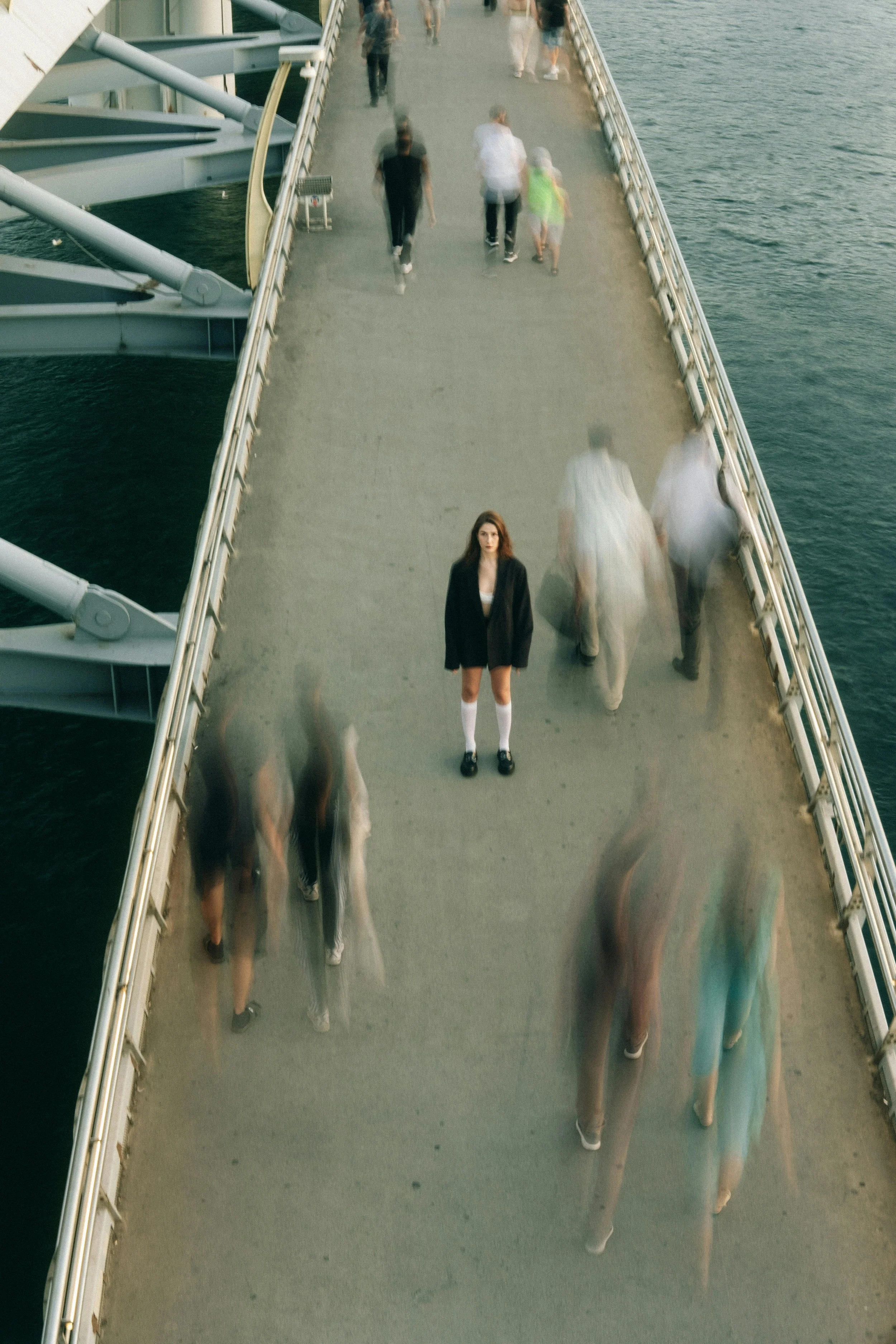 A woman stands still at the center of a pedestrian bridge, facing the camera, while blurred figures move quickly around her in both directions, creating a contrast between her stillness and the surrounding motion over the water below.