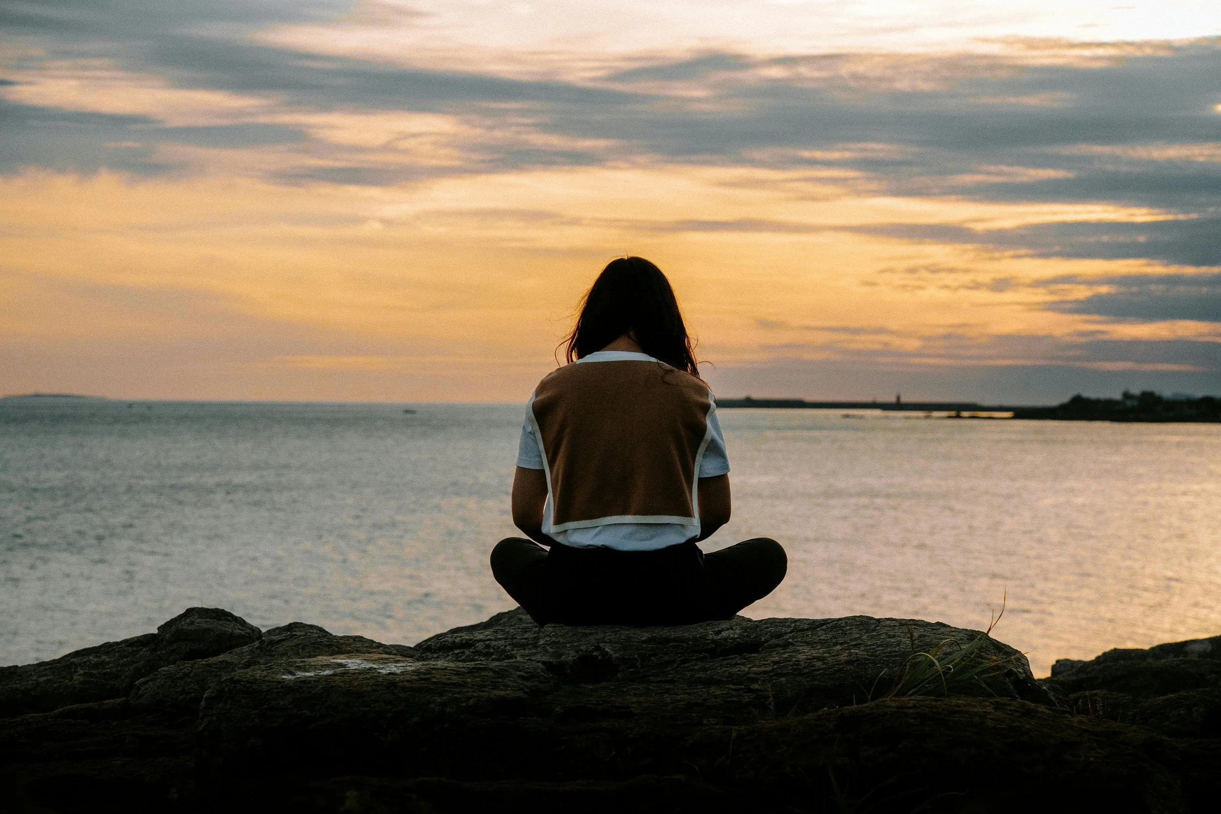 A person sits cross-legged on a rock facing a calm body of water at sunset, viewed from behind, with soft golden light reflecting on the water and clouds in the sky, conveying stillness and quiet reflection.