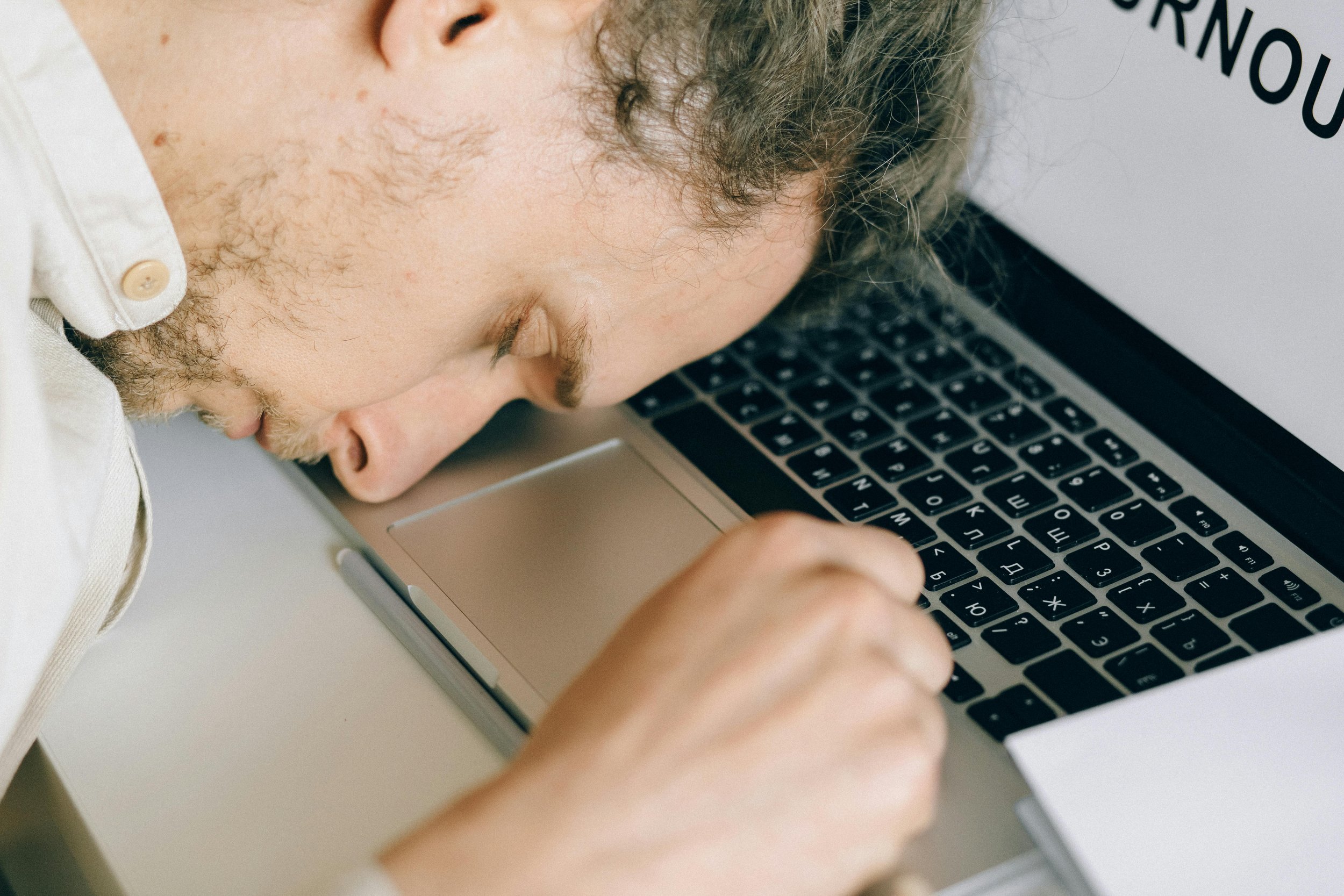 A person leaning forward with their head resting on a laptop keyboard, eyes closed, conveying tiredness while working at a desk.