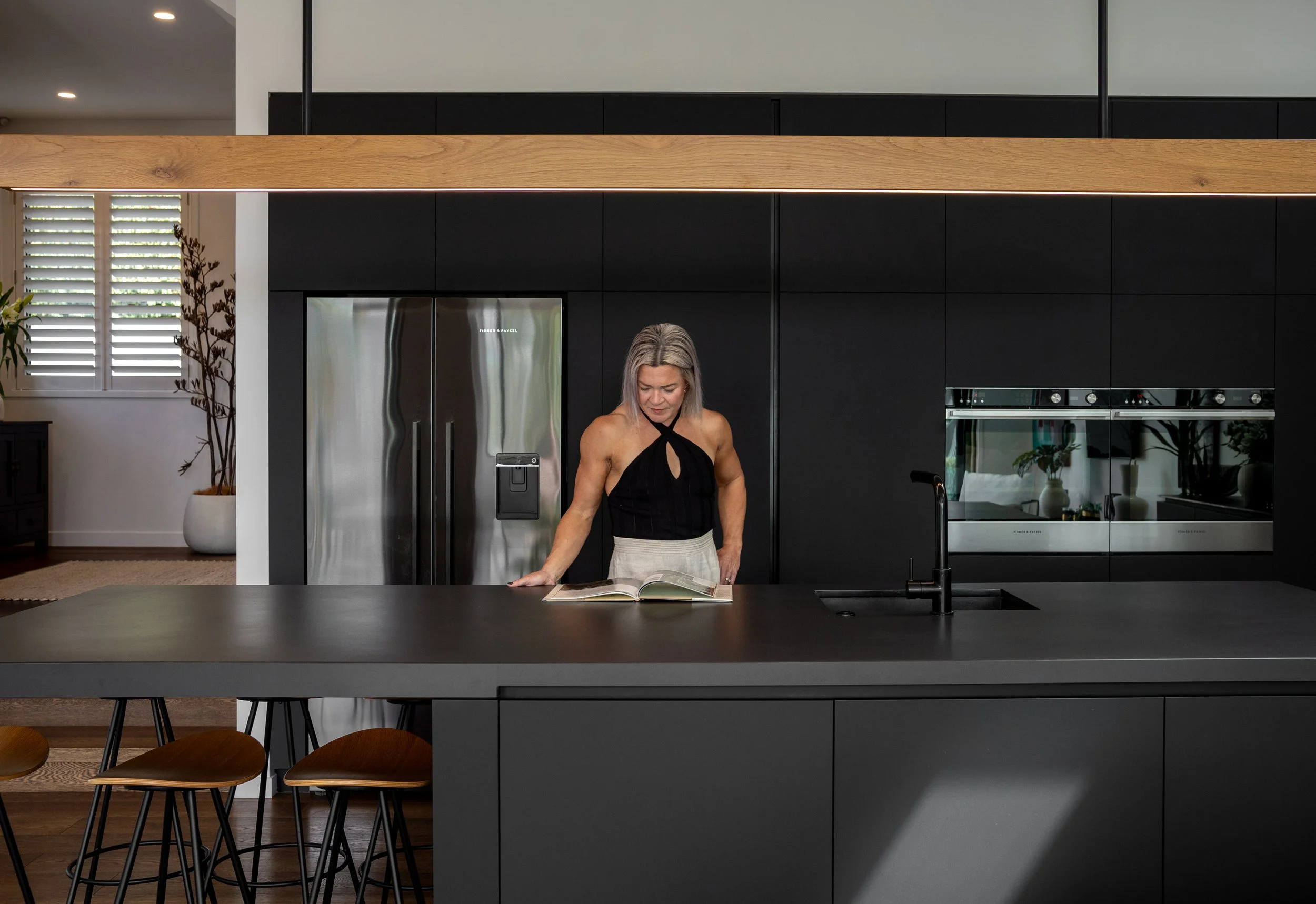 Modern kitchen interior with a person reading a cookbook on a large black island, featuring bar stools, stainless steel appliances, and sleek cabinetry.