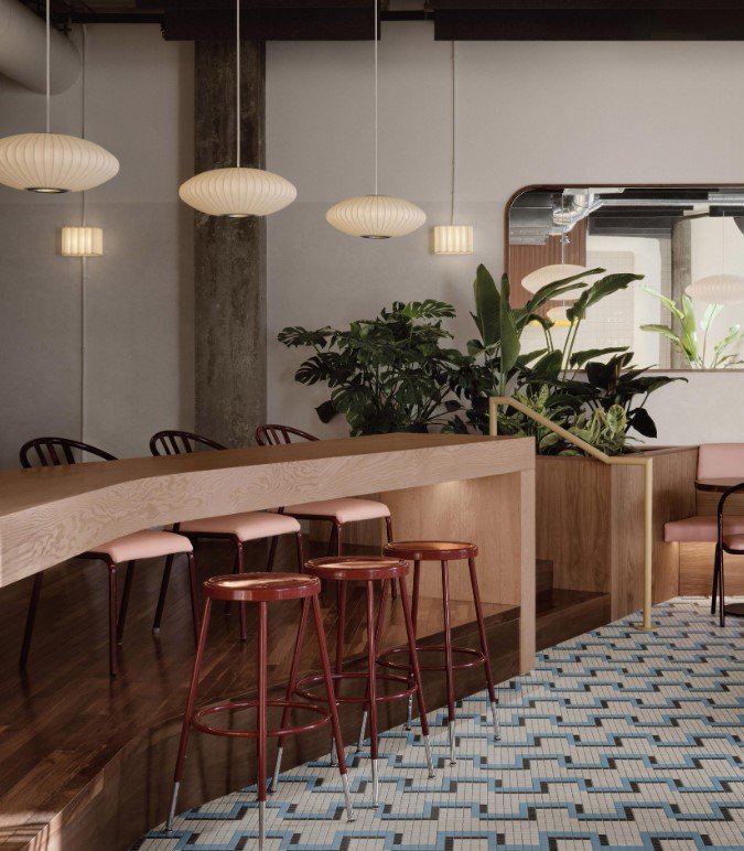 Modern cafe interior with wooden bar, red stools, hanging pendant lights, and green plants. The floor has a patterned blue and white tile design.