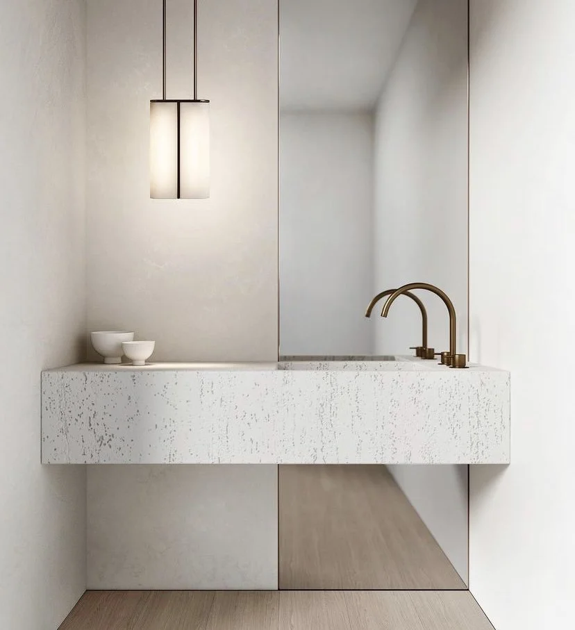 Minimalist bathroom with modern pendant light, textured white countertop, twin brass faucets, and two small white bowls.