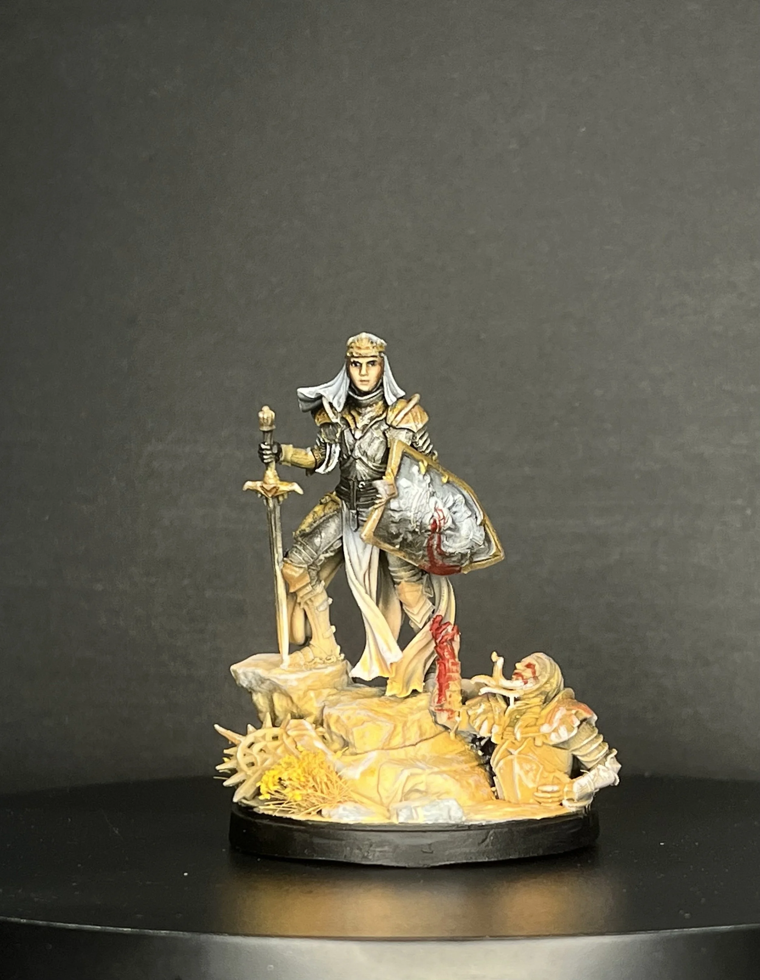 A detailed miniature figure of a female warrior with a sword and shield, standing on a rocky base with battle damage and fallen armor.