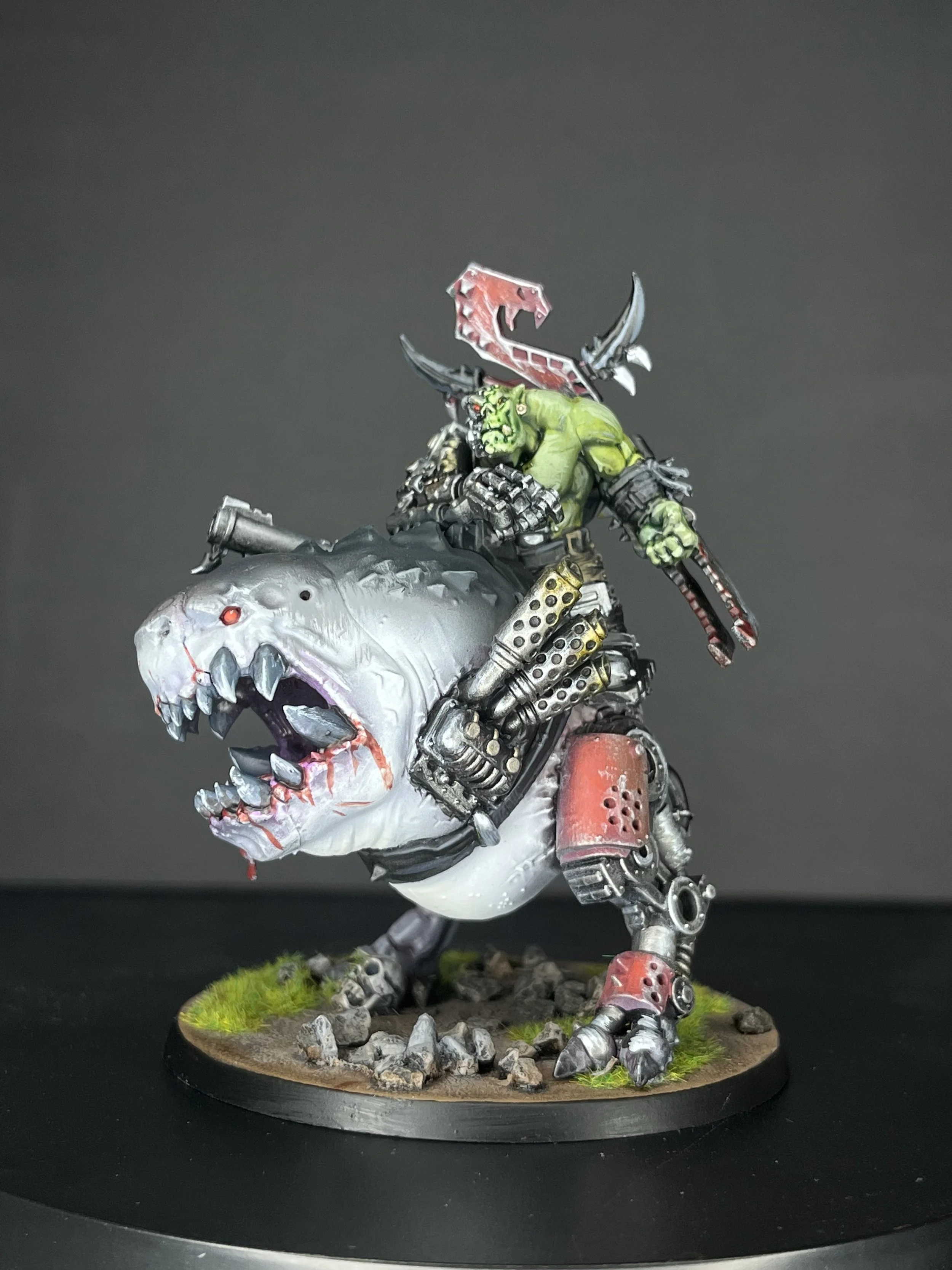 Miniature model of a green orc riding a robotic dinosaur with a large head and open mouth, all painted with detailed weathering and standing on a textured base with rocks and grass.