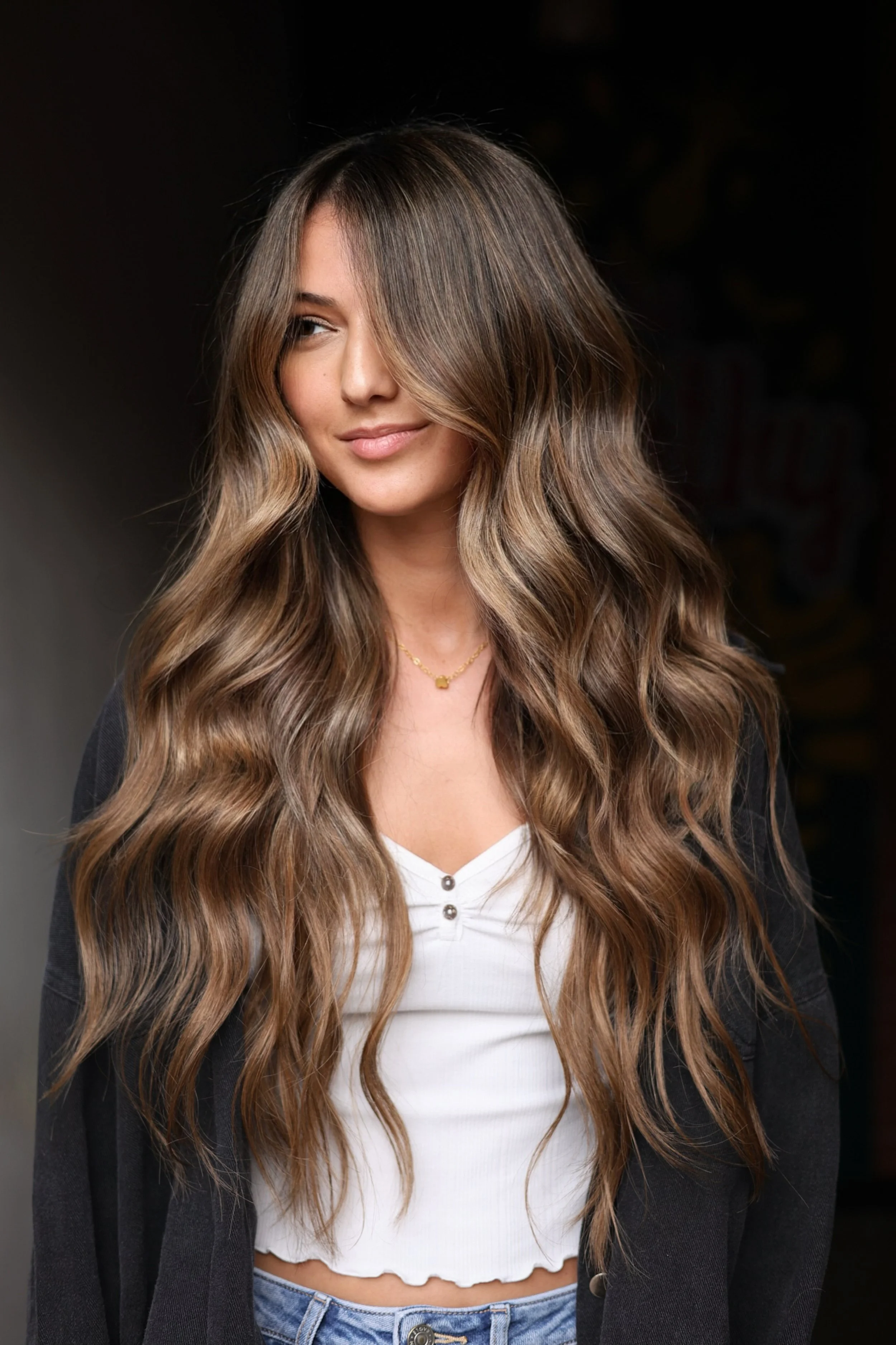 A young woman with long wavy light brown hair, partially covering her face with a lock, wearing a black jacket over a white button-up top and a delicate gold necklace, standing against a dark background.