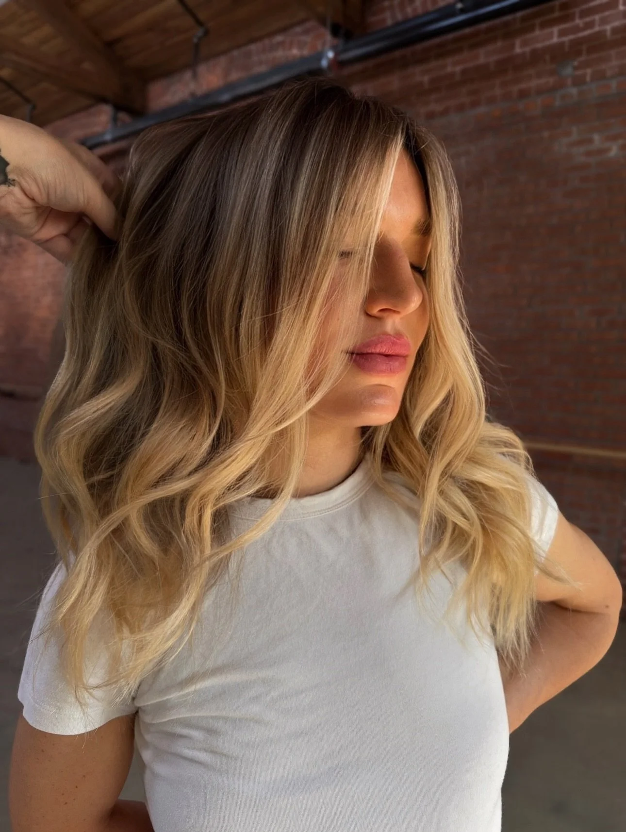 A woman with long blonde hair, wearing a white t-shirt, standing with her eyes closed and a hand in her hair inside a brick-walled room.