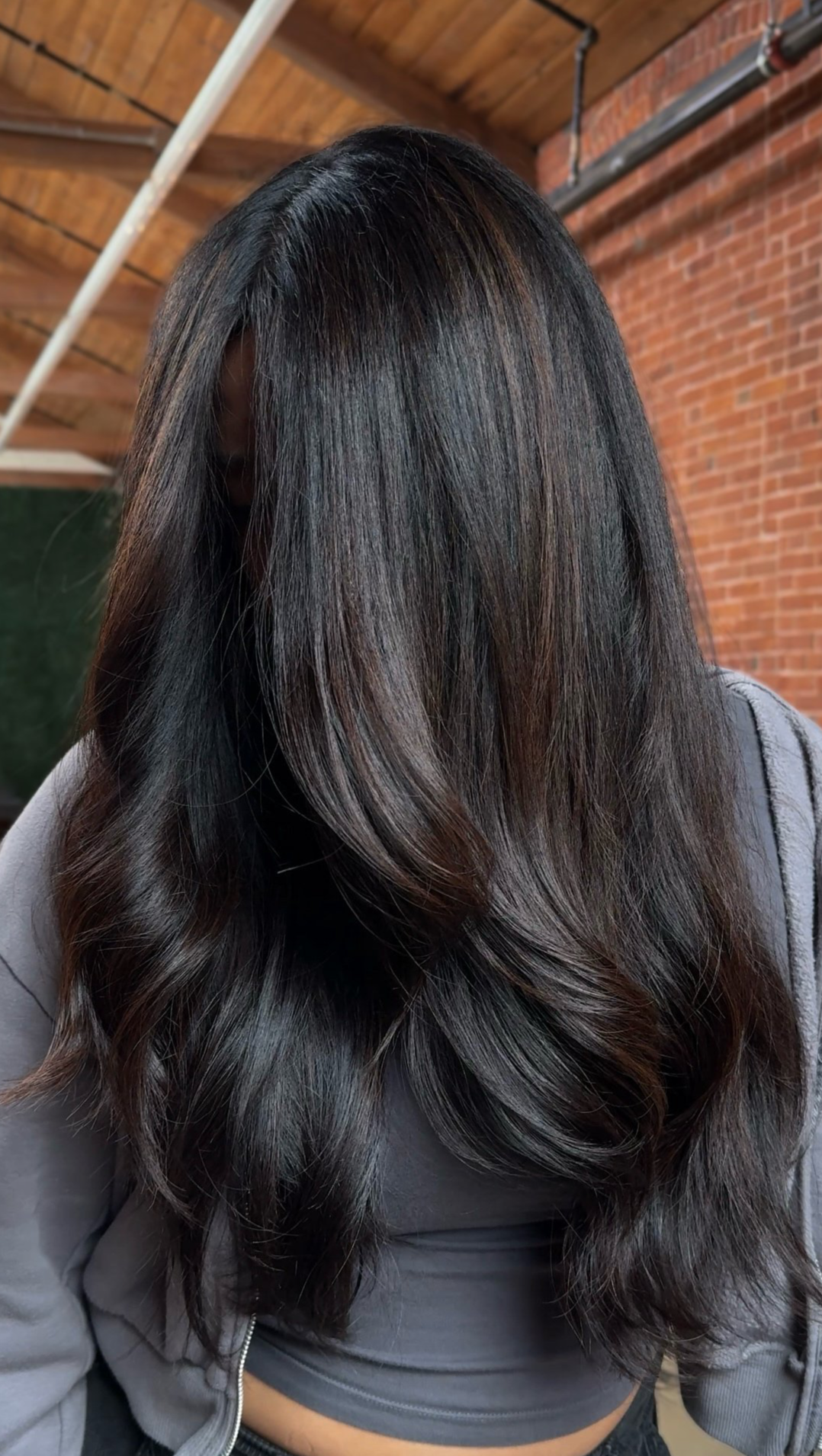 Close-up of a woman with long, shiny, dark brown hair with subtle lighter highlights, indoors with a brick wall and wooden ceiling in the background.
