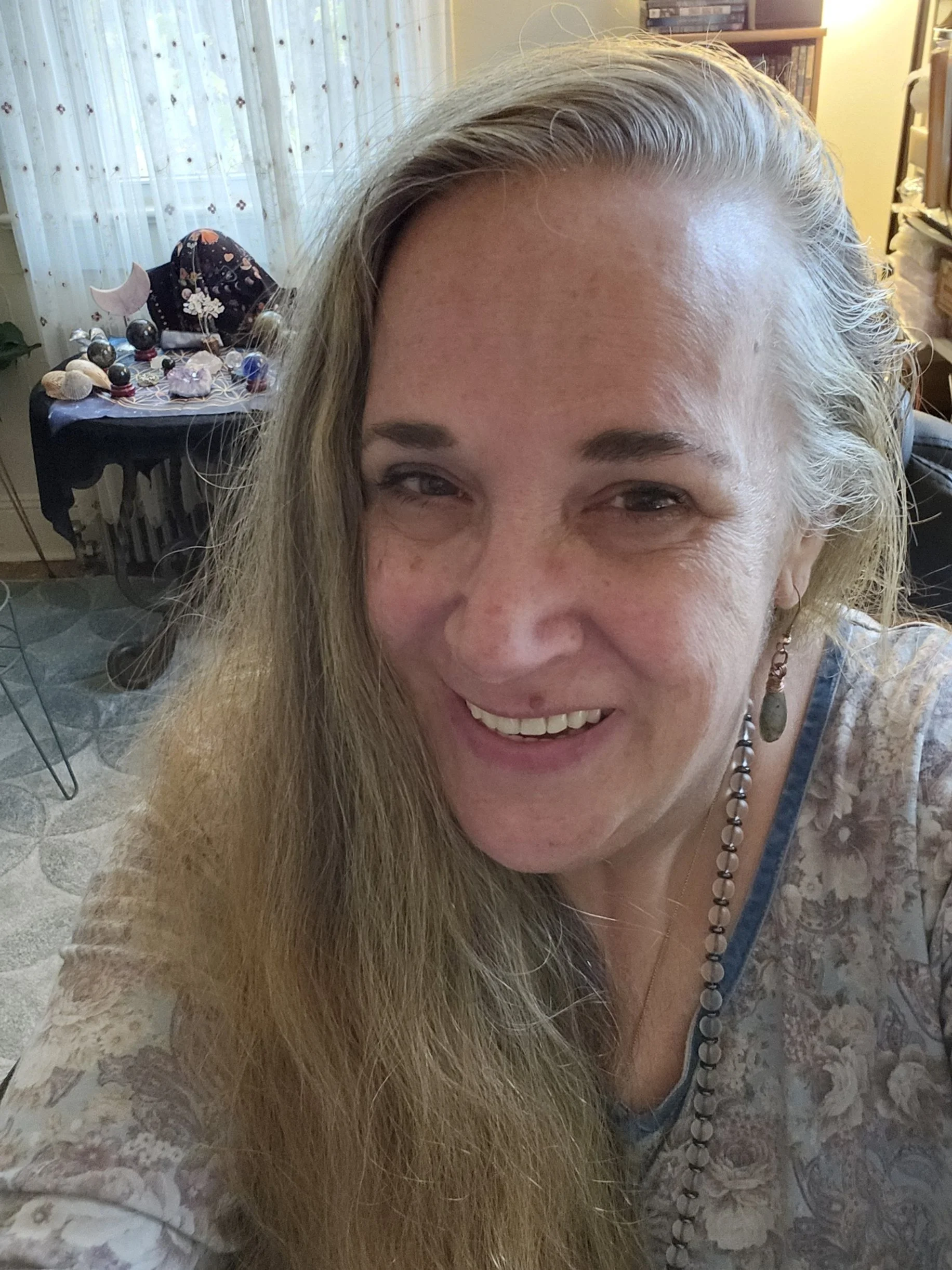 A smiling woman with long blonde hair and a beaded necklace in a cozy room with a decorated table in the background.