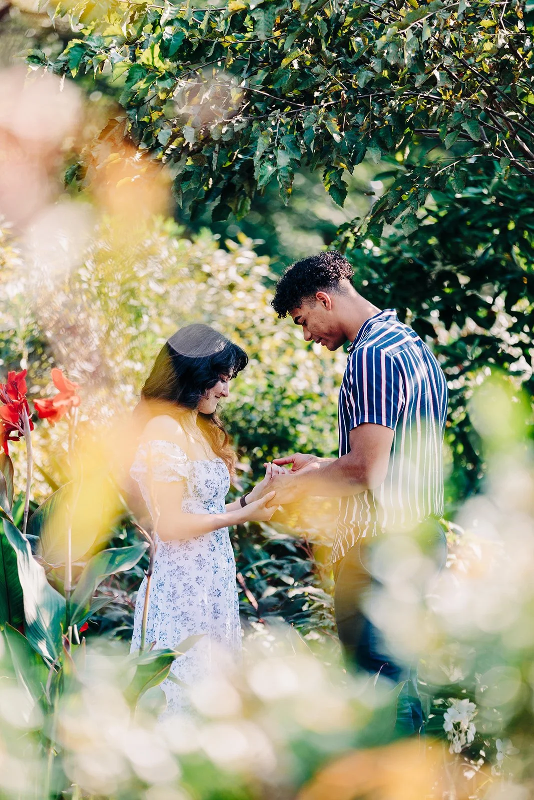 A couple exchanging rings in a lush, green outdoor setting with flowering plants. The woman is wearing a white, floral dress, and the man is dressed in a striped shirt.