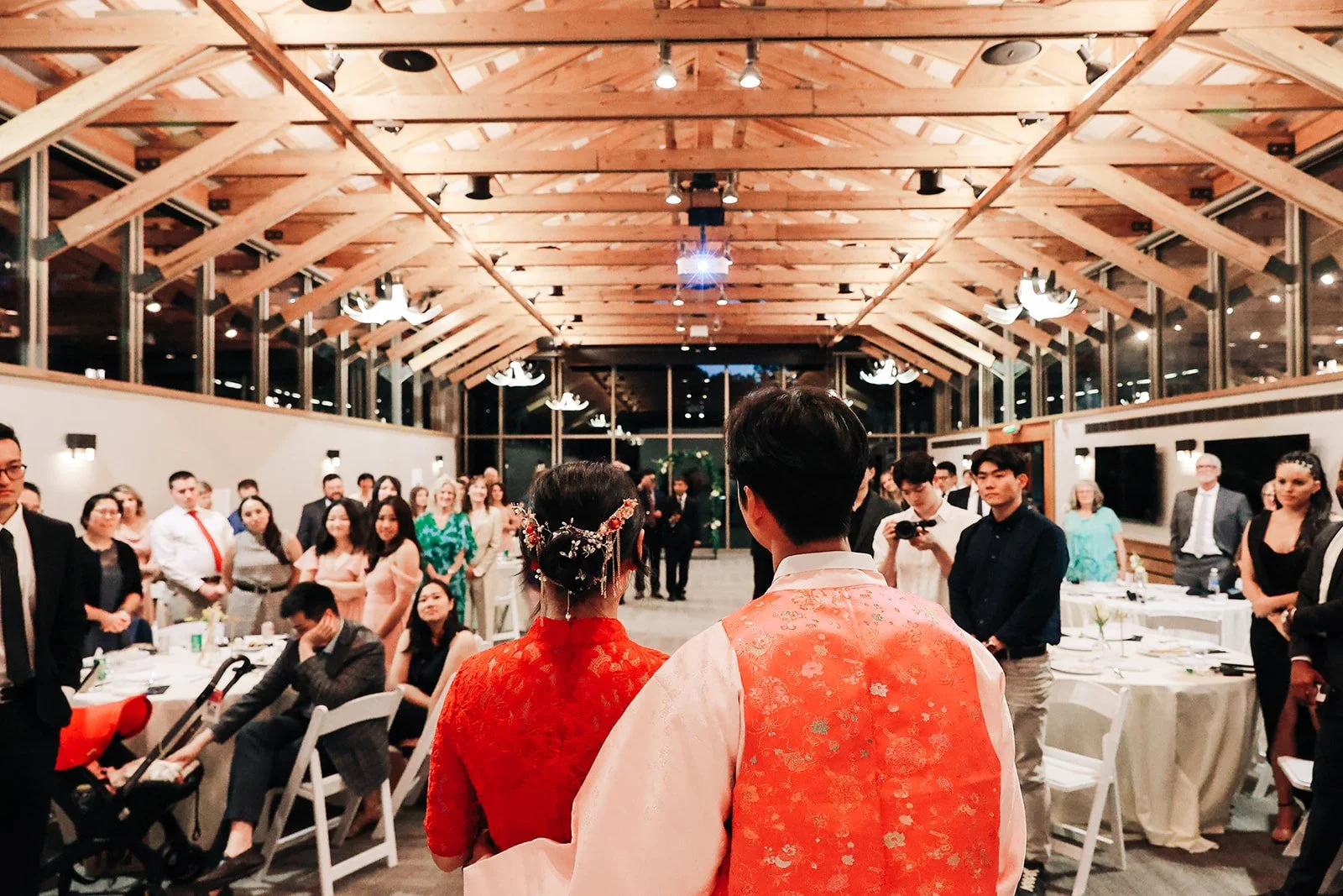 Couple in traditional Asian attire at a wedding reception, facing a large group of guests in a warmly lit, modern event hall with wooden ceiling, large windows, and elegant chandeliers.