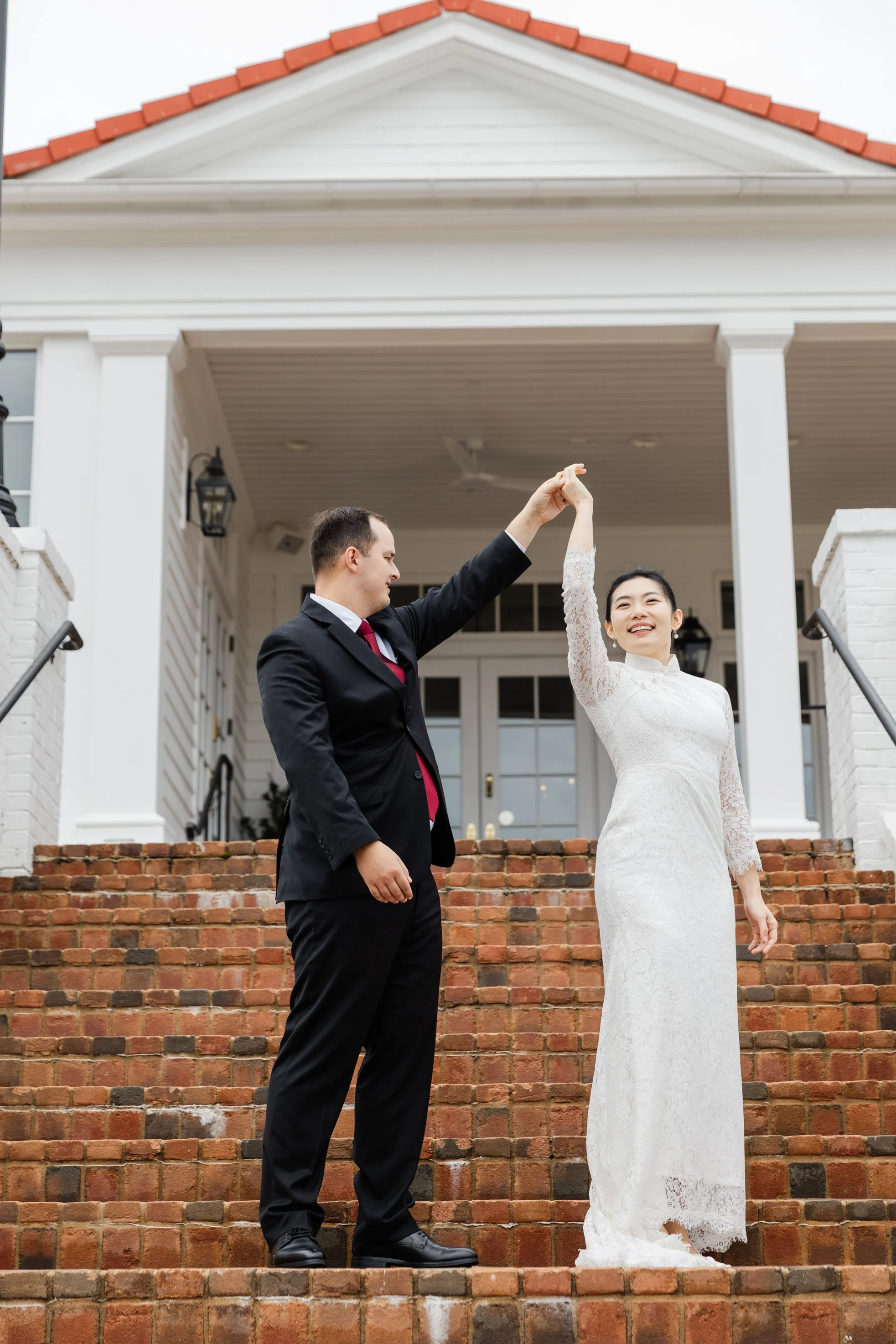 A couple dancing on brick stairs outside a white house, with the man in a black suit and the woman in a white lace dress, smiling and holding hands above their heads.