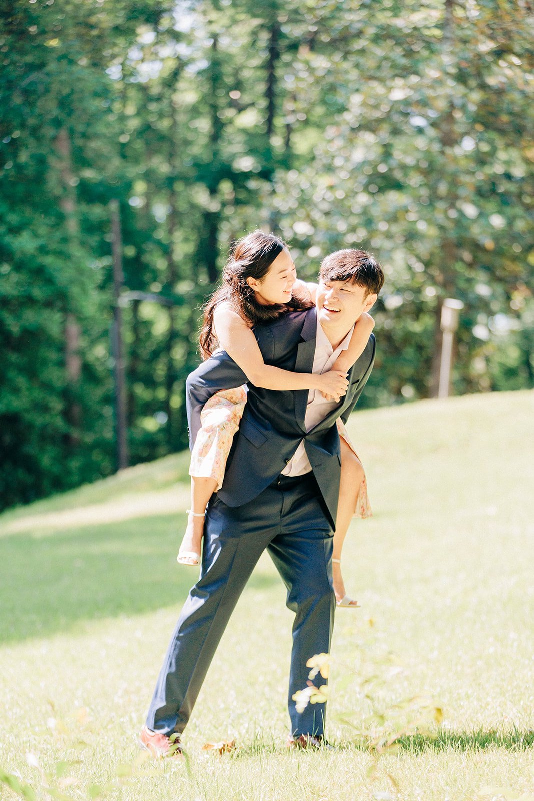 A young man in a suit giving a piggyback ride to a young woman in a park with green trees, both smiling and enjoying the sunny day.