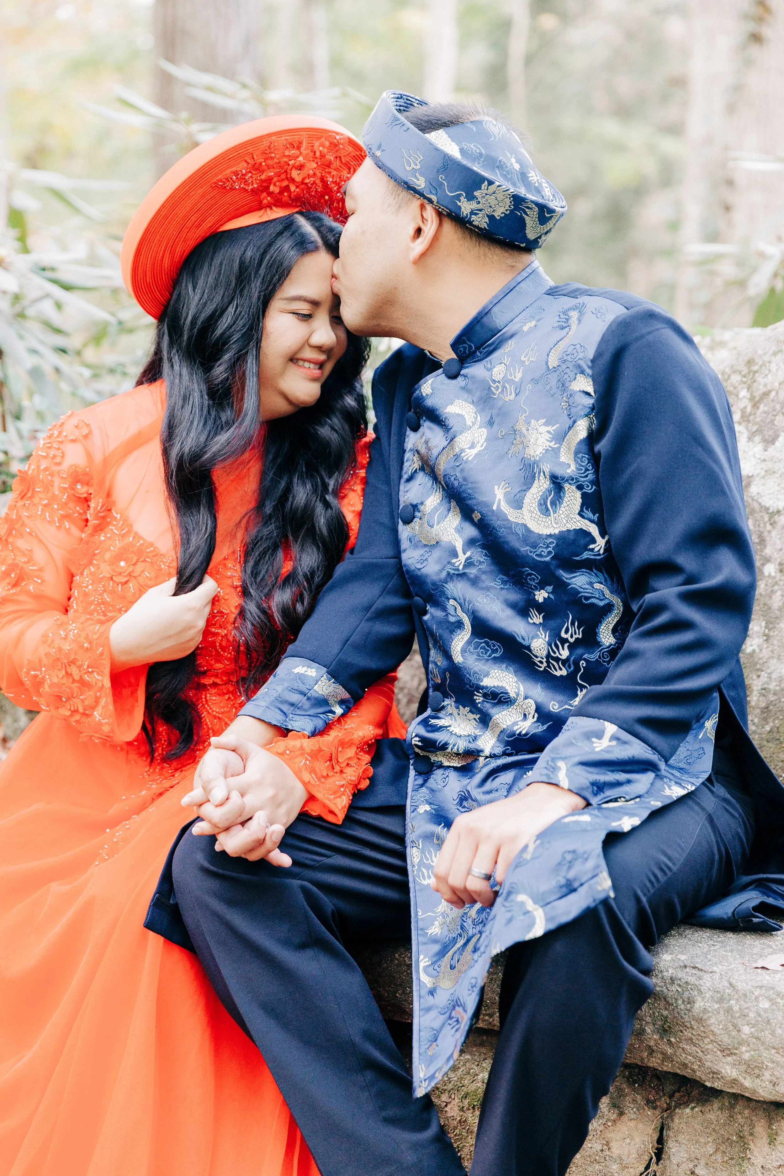 A couple dressed in traditional Vietnamese attire, with the woman in an orange ao dai and the man in a blue áo dài, sitting outdoors on a rock, sharing a tender moment with the man kissing the woman's forehead.