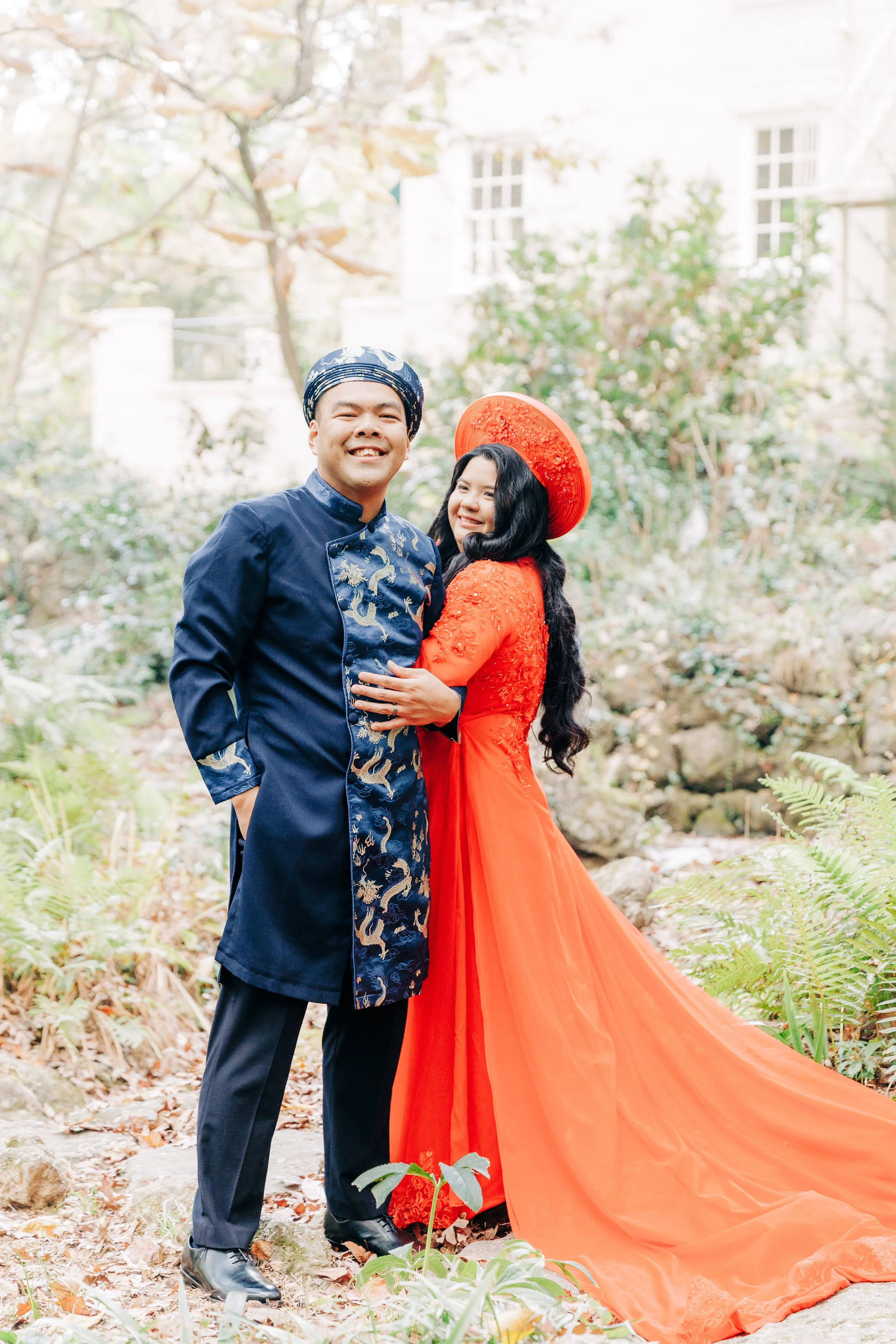 A couple dressed in traditional Vietnamese clothing standing outdoors in a garden with autumn leaves and trees. The man is wearing a navy blue outfit with gold embroidery and a matching hat. The woman is wearing a bright orange ao dai and a matching 