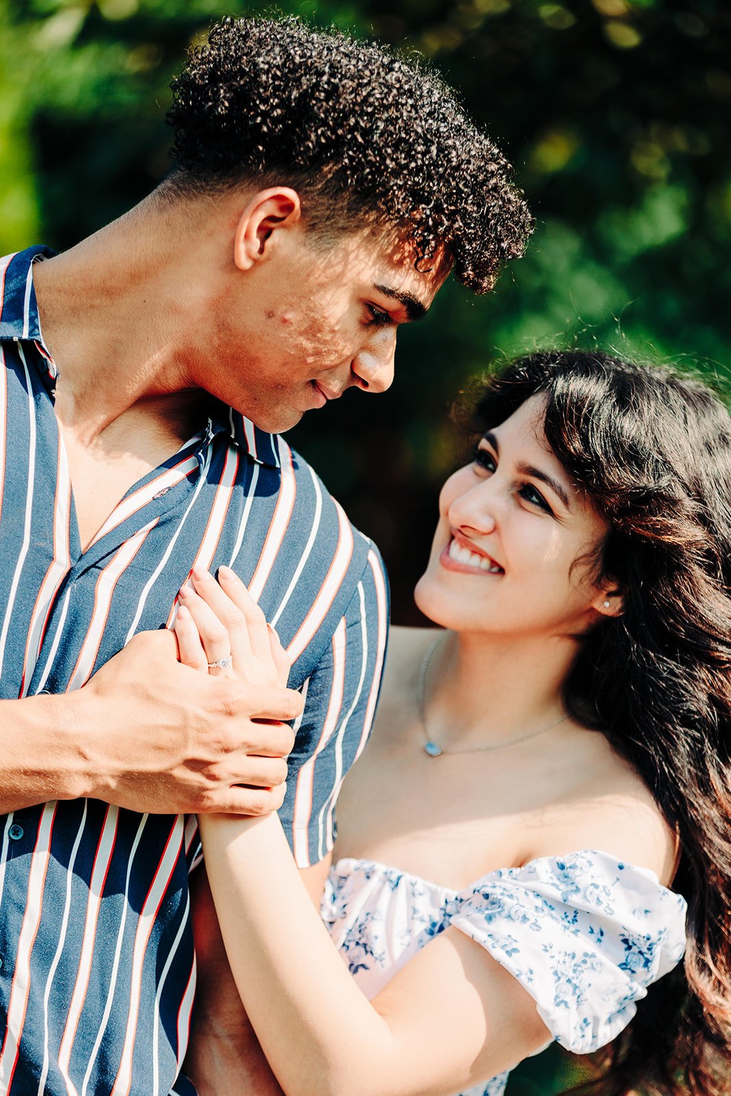 A young couple smiling and looking at each other outdoors with a blurred green background. The woman is wearing a white off-shoulder dress and a necklace, while the man is wearing a blue striped shirt. The woman has her hand on the man's shoulder, sh