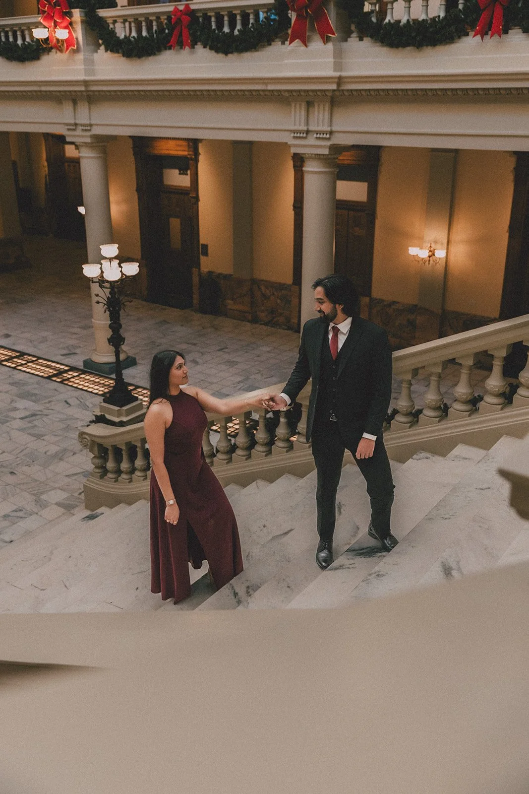 A man in a dark suit and red tie helps a woman in a long burgundy gown on a staircase inside a decorated building with Christmas garlands and ribbons.