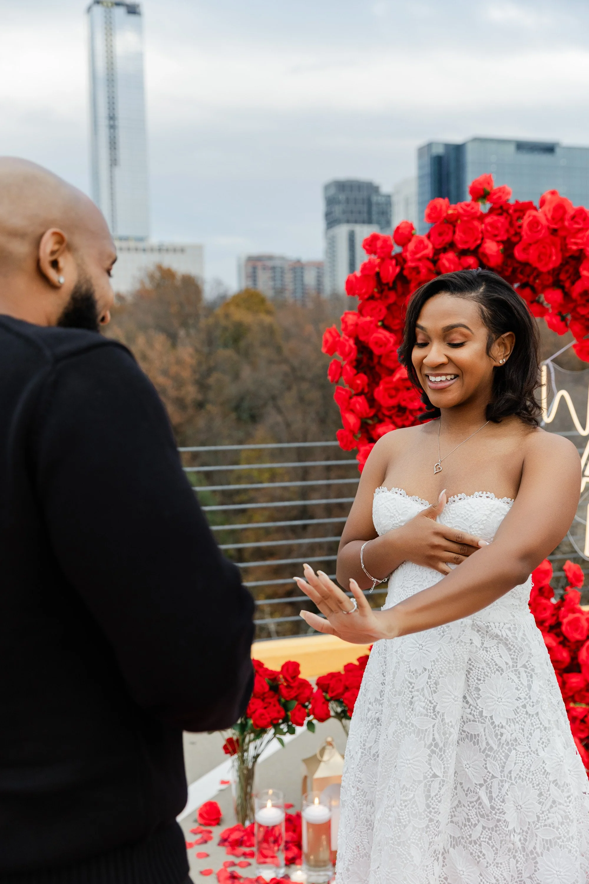 A woman in a white lace strapless dress happily saying no during a wedding proposal with a man in a black sweater, surrounded by a floral display of red roses and candles, on a rooftop with city buildings in the background.